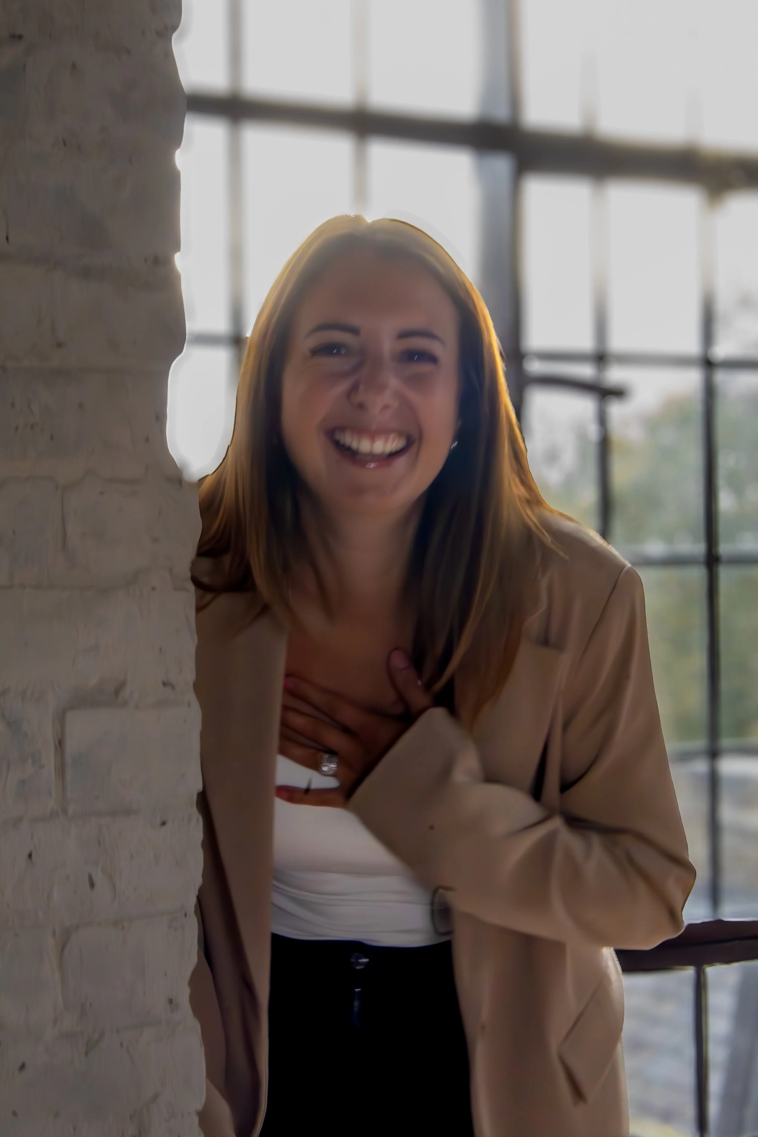 A woman with red hair, smiling, standing near a brick wall and an industrial-style window, wearing a beige blazer and a white top.