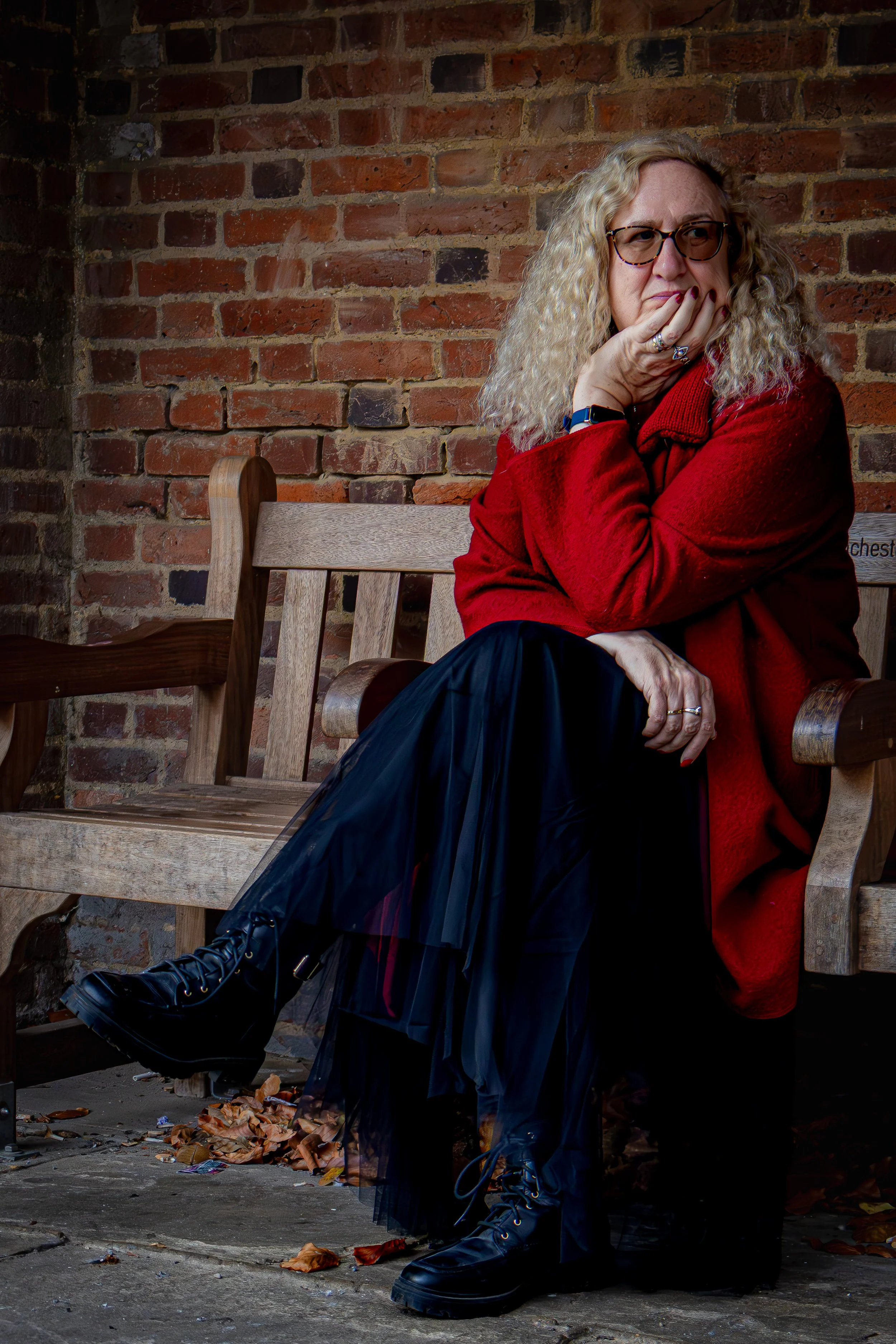 A woman with curly blonde hair, wearing glasses, a red coat, and black boots, sitting on a wooden bench against a brick wall, with her hand on her chin and a thoughtful expression.