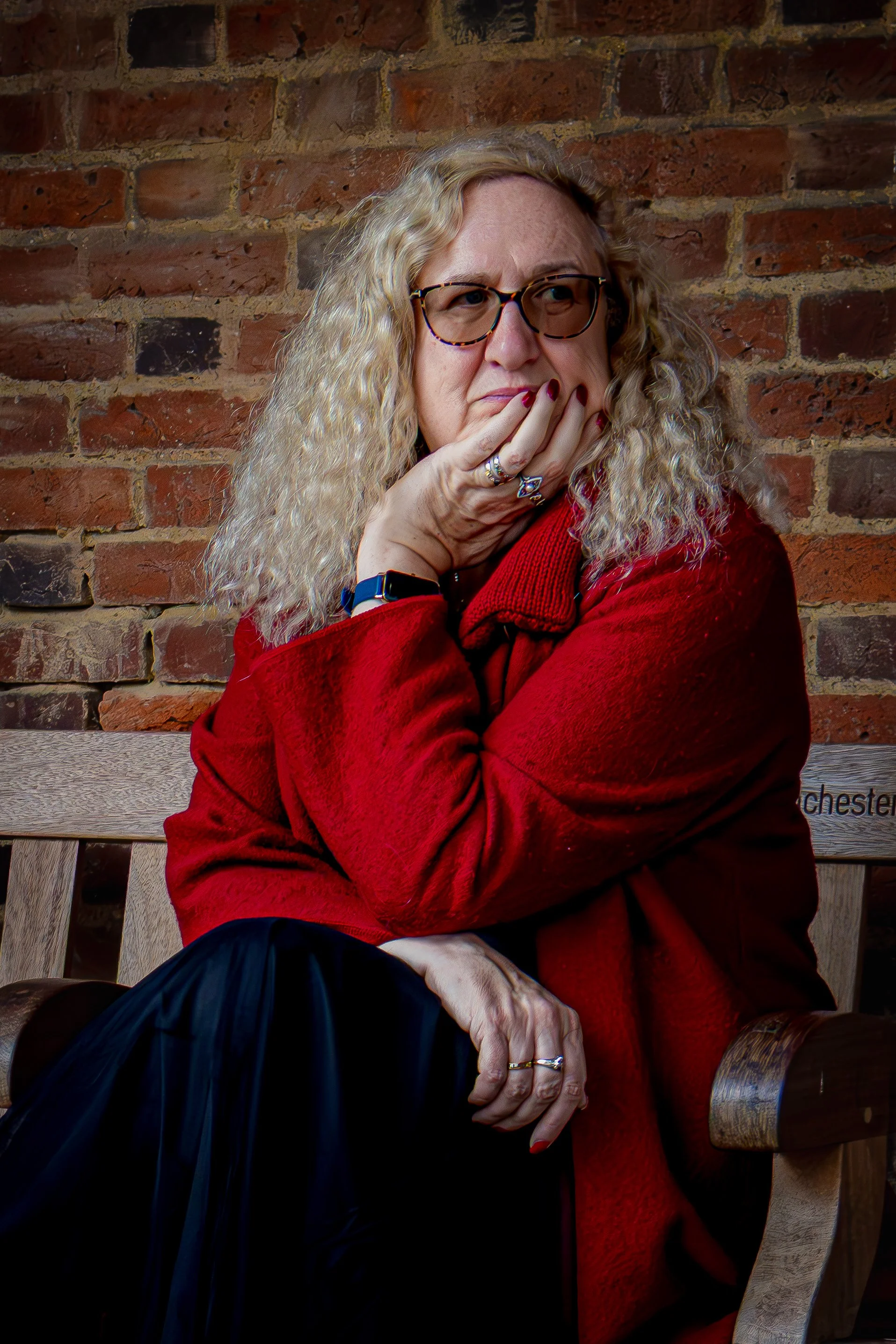 A woman with curly blonde hair and glasses sits on a wooden bench against a brick wall, wearing a red jacket, appearing thoughtful with her hand touching her face.