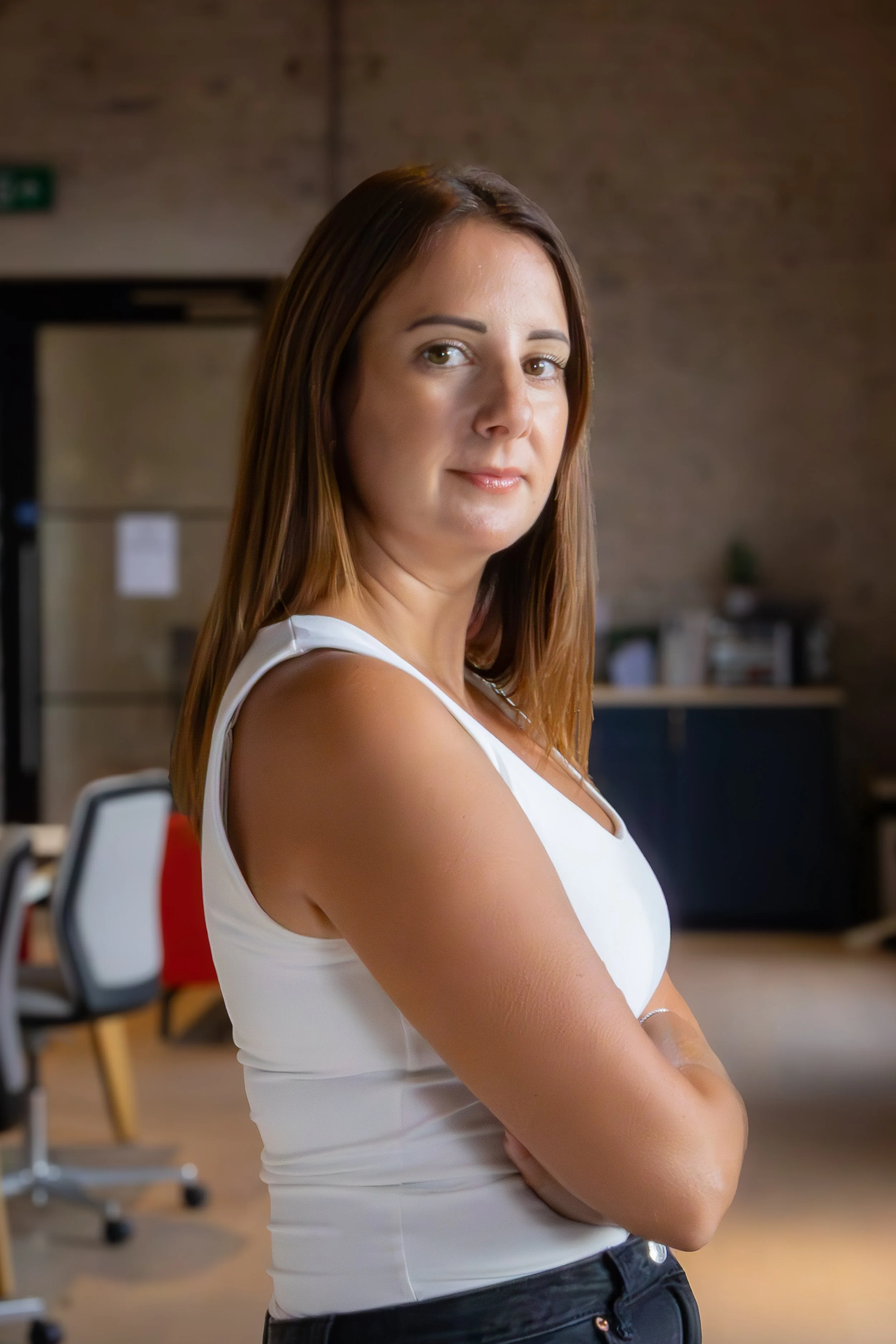 A woman with shoulder-length brown hair, wearing a white sleeveless top, standing in an office with arms crossed, looking at the camera.