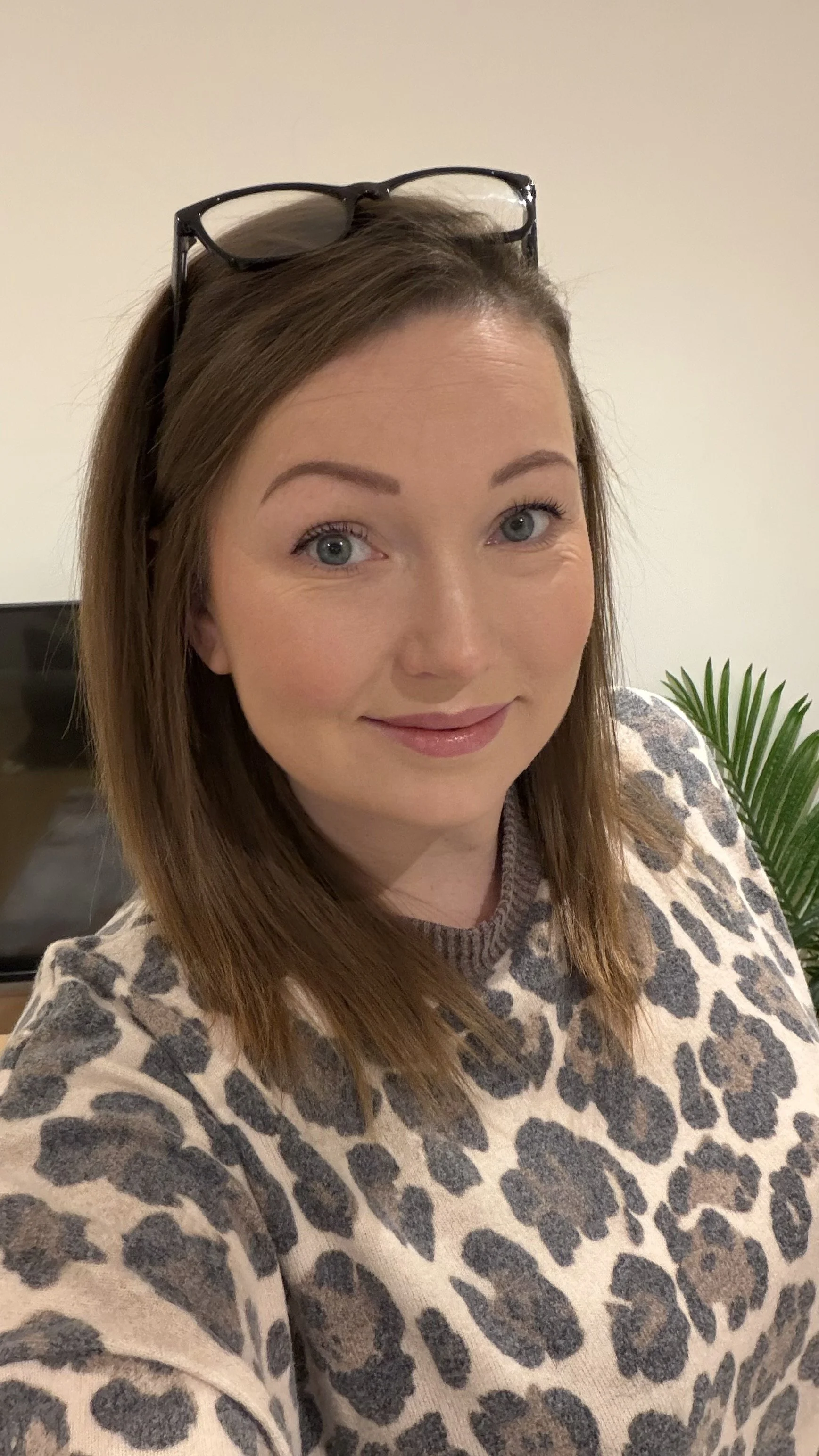 A woman with brown hair wearing glasses on her head and a leopard print sweater, smiling in front of a plain wall and a potted plant. This is Dani, the founder of Build the Buzz Socials and Photos.