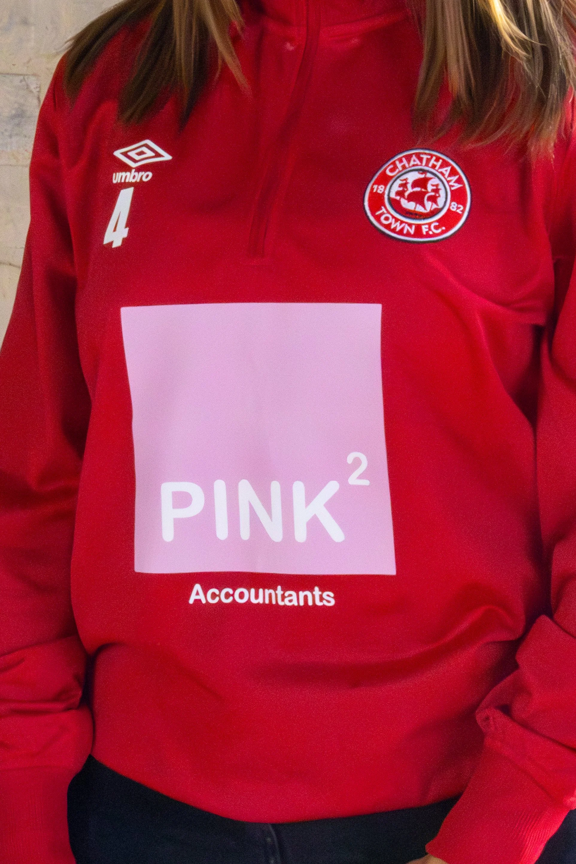 A person wearing a red sports jacket with the Chatham Town Football Club logo on the left chest, the Umbro logo and number 4 on the right chest, and a pink sponsor logo with the text 'PINK² Accountants' on the front.