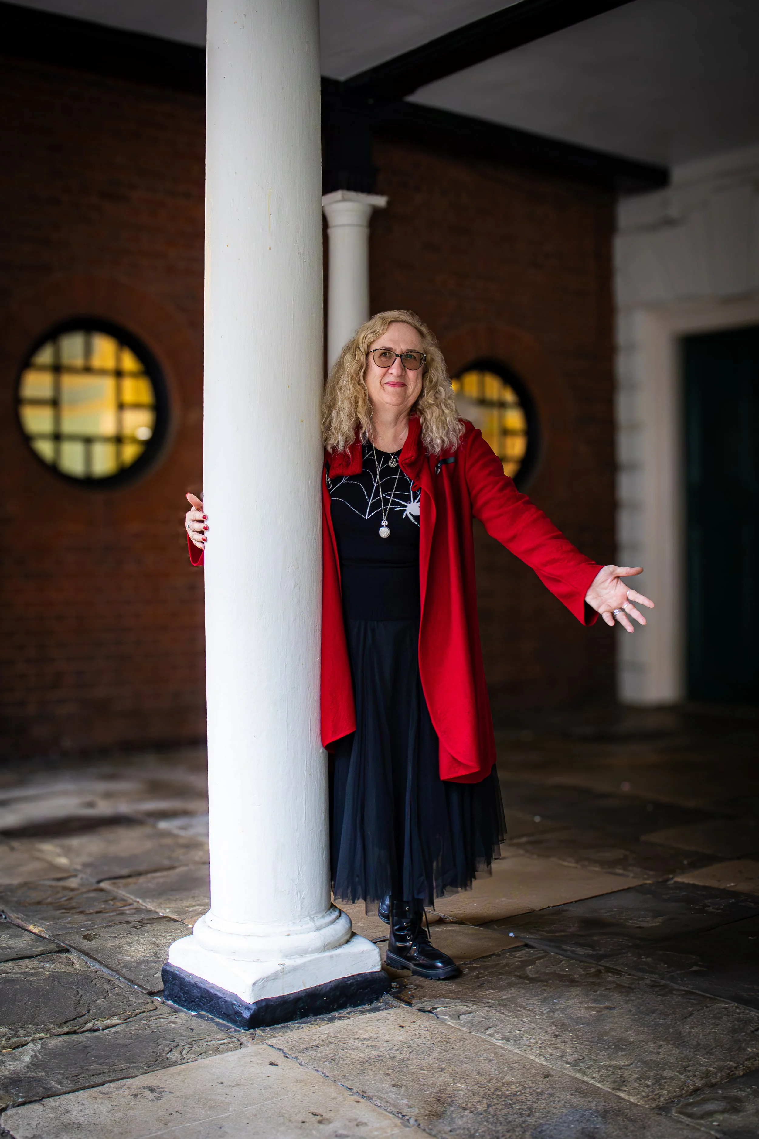 A woman with curly blonde hair, wearing glasses, a black dress, a red coat, and black boots, standing in front of a white column on a stone pavement at night.