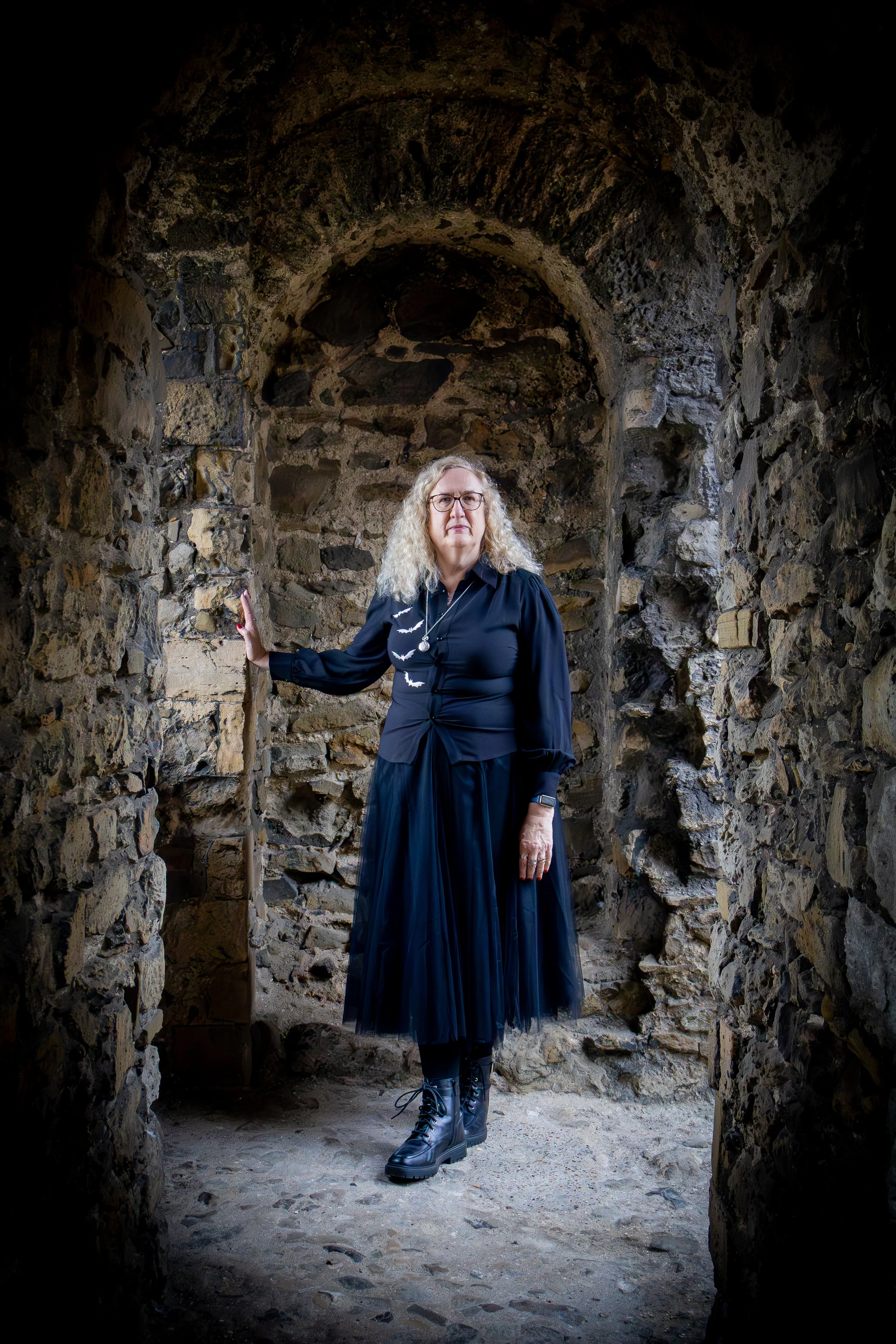 A woman with curly blonde hair and glasses standing inside a stone archway, dressed in a black shirt, black skirt, and combat boots.