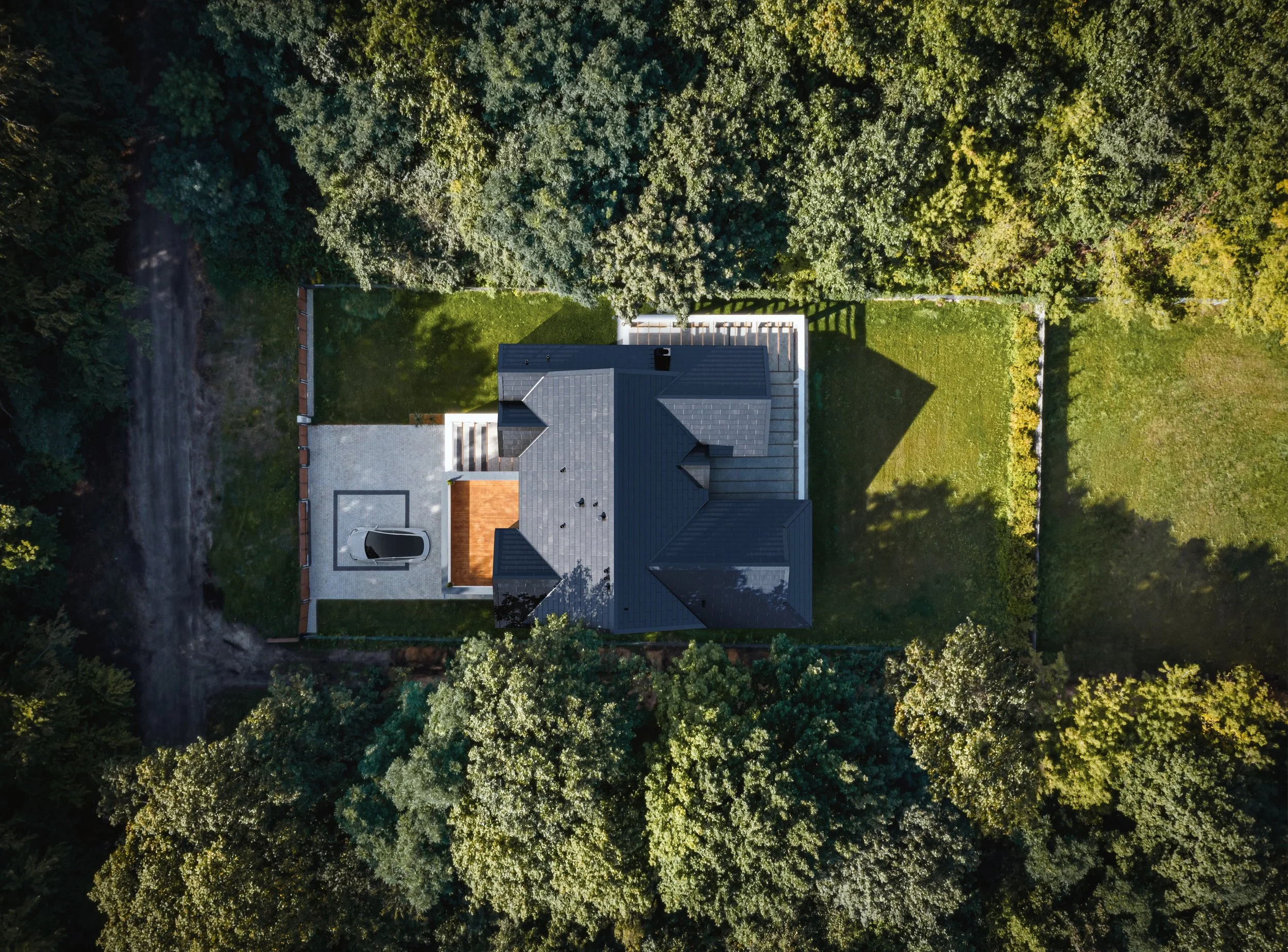 Aerial view of a house with a dark gray roof, surrounded by green trees and lawns, with a driveway and a parked car in the front yard.