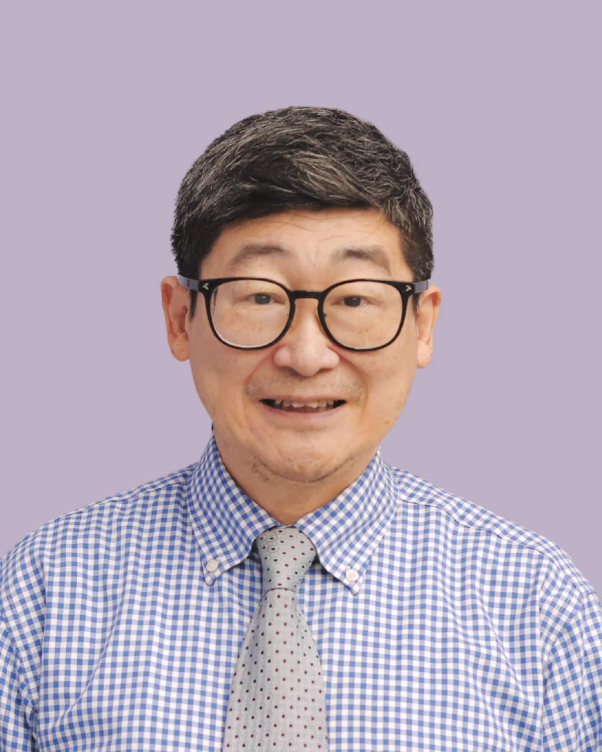A middle-aged man with black hair, glasses, wearing a blue and white checkered shirt and a light gray tie with small red and black dots, smiling against a plain light purple background.