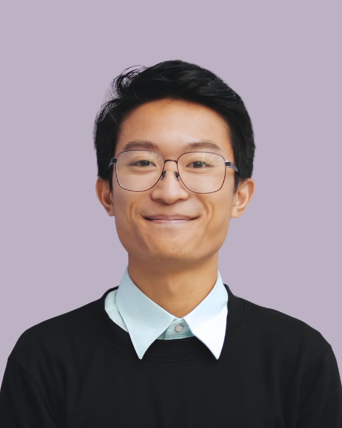 A young man smiling, wearing glasses, a light blue collared shirt, and a black sweater, against a purple background.