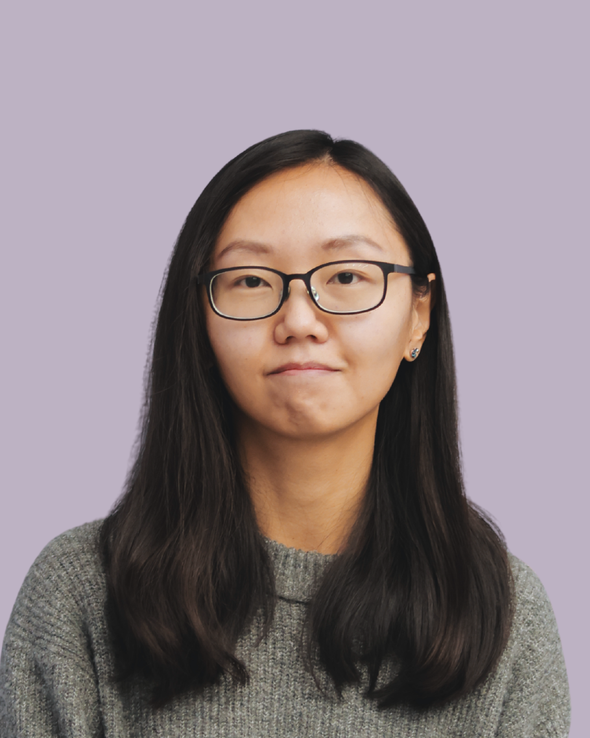 Portrait of a young woman with long dark hair wearing glasses and a gray sweater, against a light purple background.