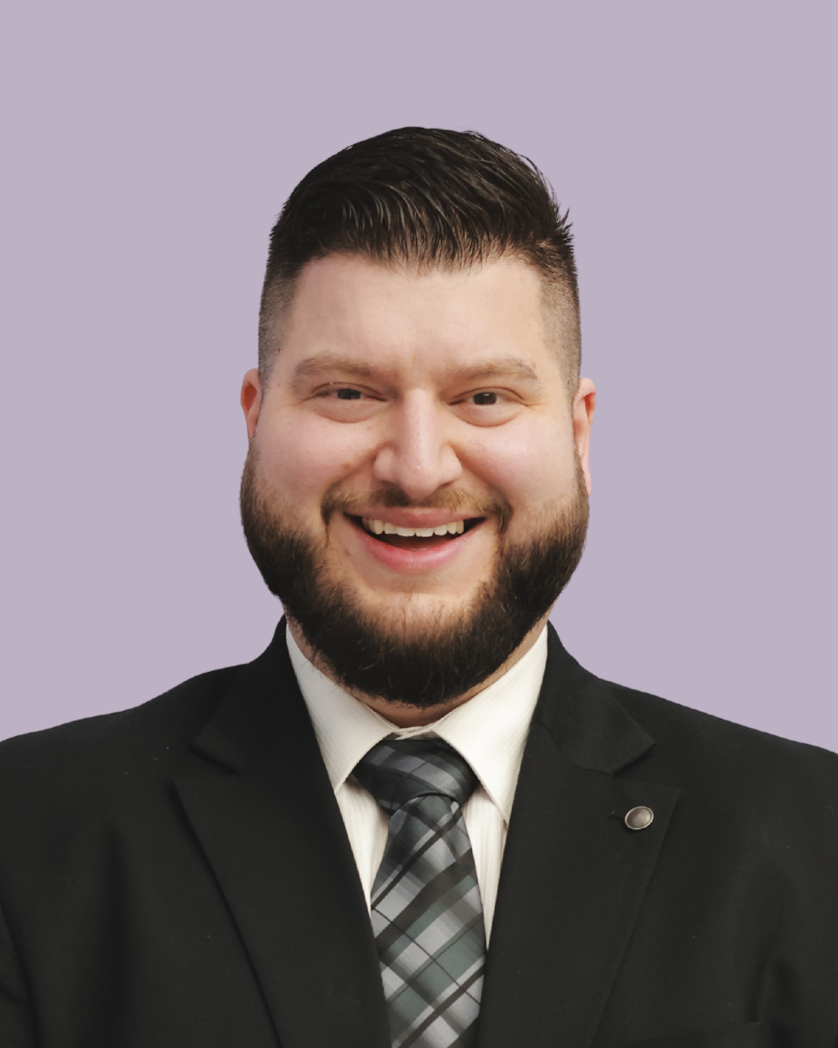 Headshot of a smiling man with dark hair and beard, wearing a black suit, white shirt, and plaid tie, against a light purple background.