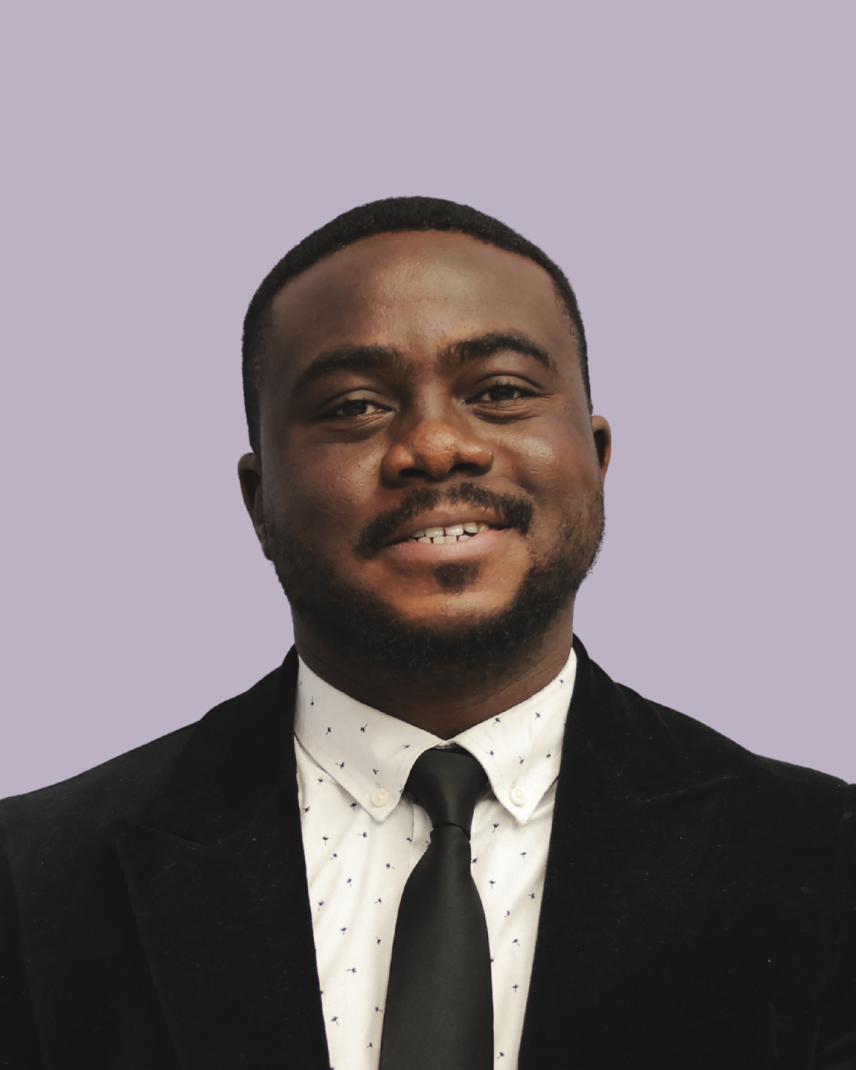 A man with dark skin, short black hair, and a beard, dressed in a black suit jacket, white shirt with small dark patterns, and a black tie, standing against a light purple background.