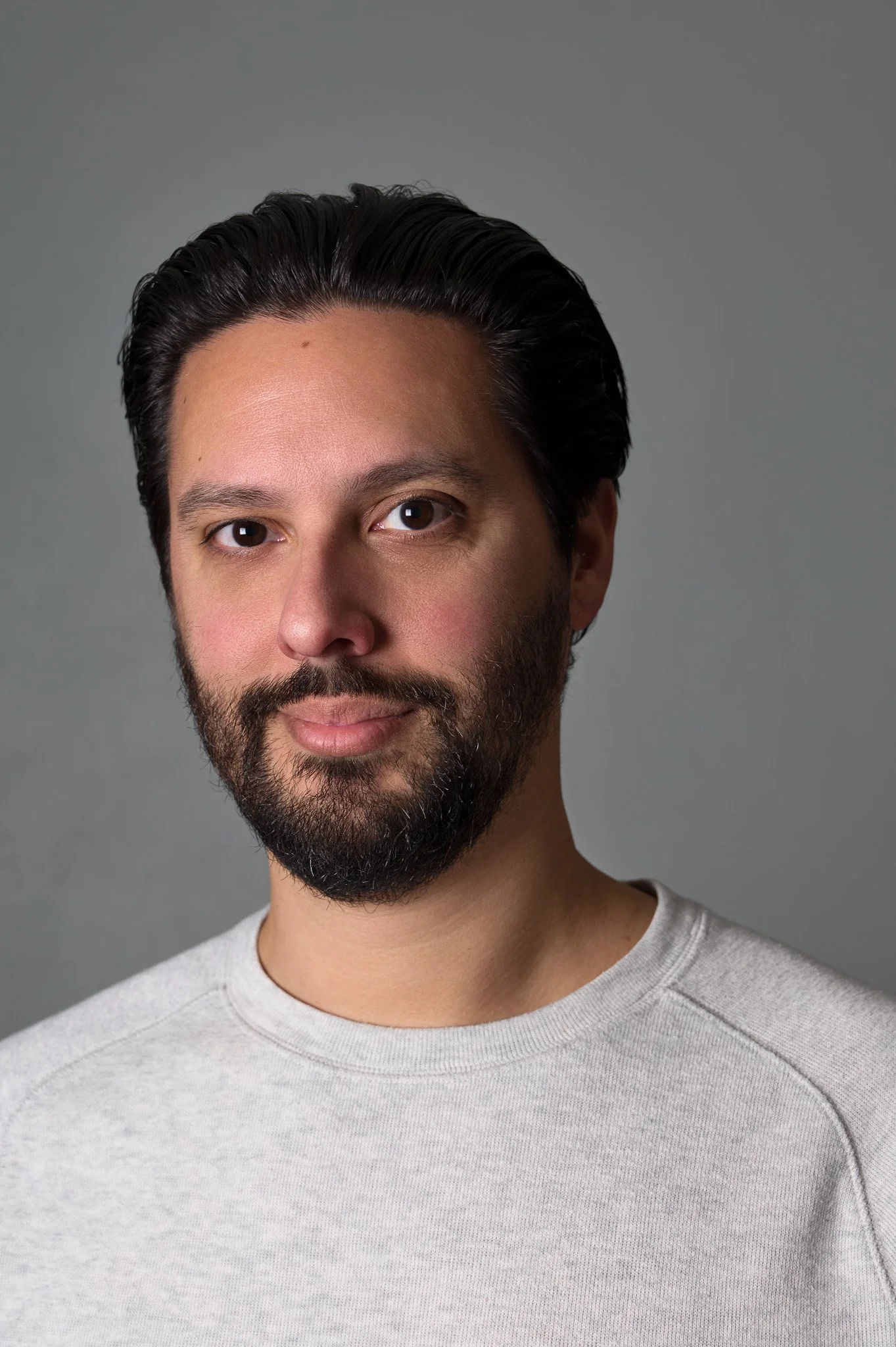 Close-up of a man with dark hair and beard, wearing a light grey t-shirt, against a grey background.