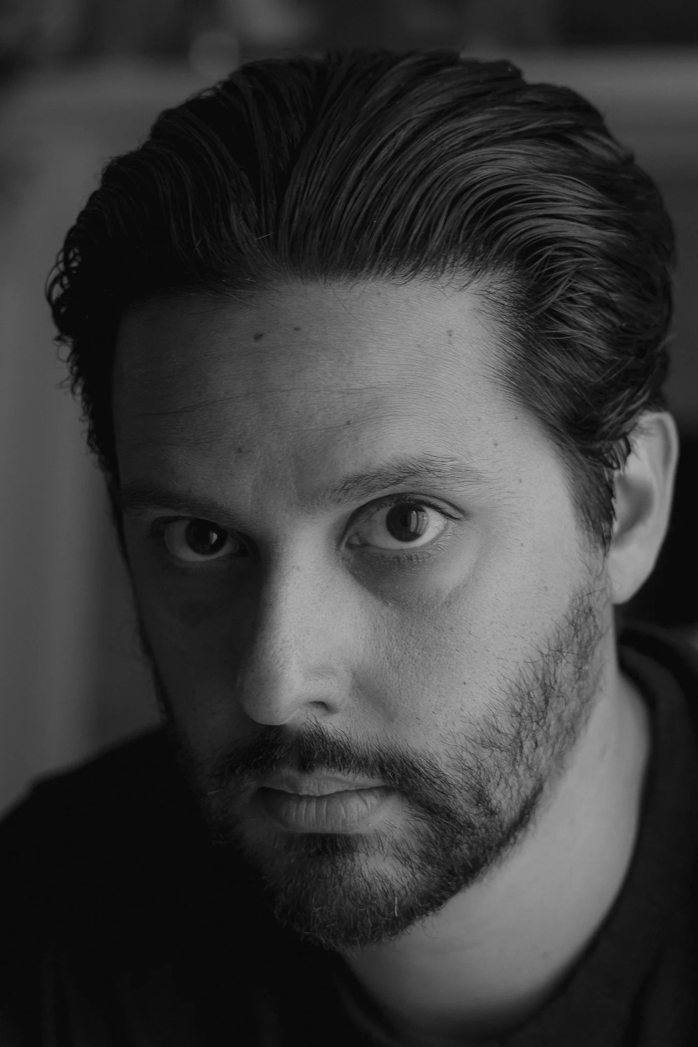 Close-up black and white portrait of a man with slicked-back hair and a beard, looking directly at the camera.