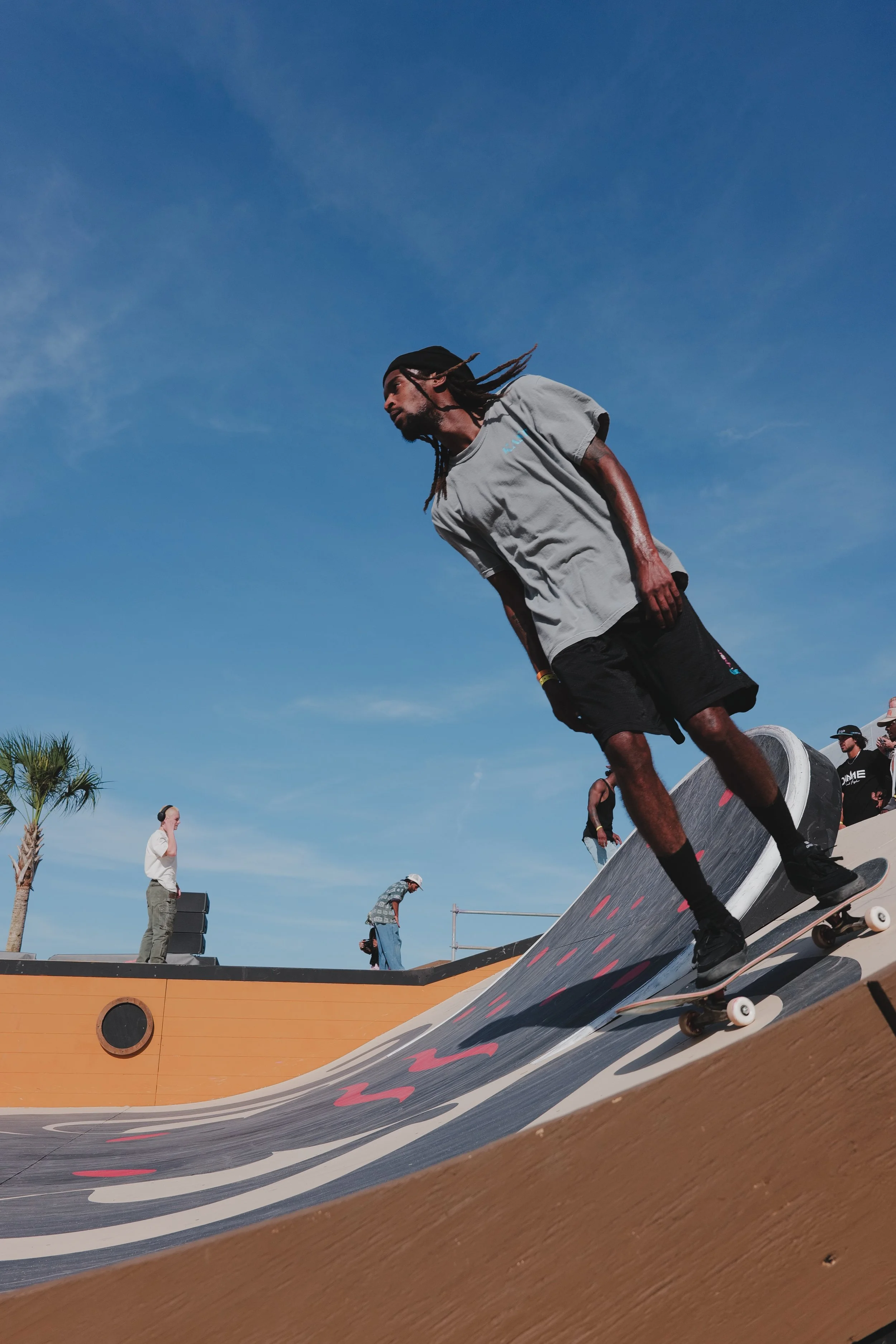 A skateboarder wearing a gray shirt and black shorts rides on a curved ramp under a clear blue sky. Other people are visible in the background near the ramp and palm trees.