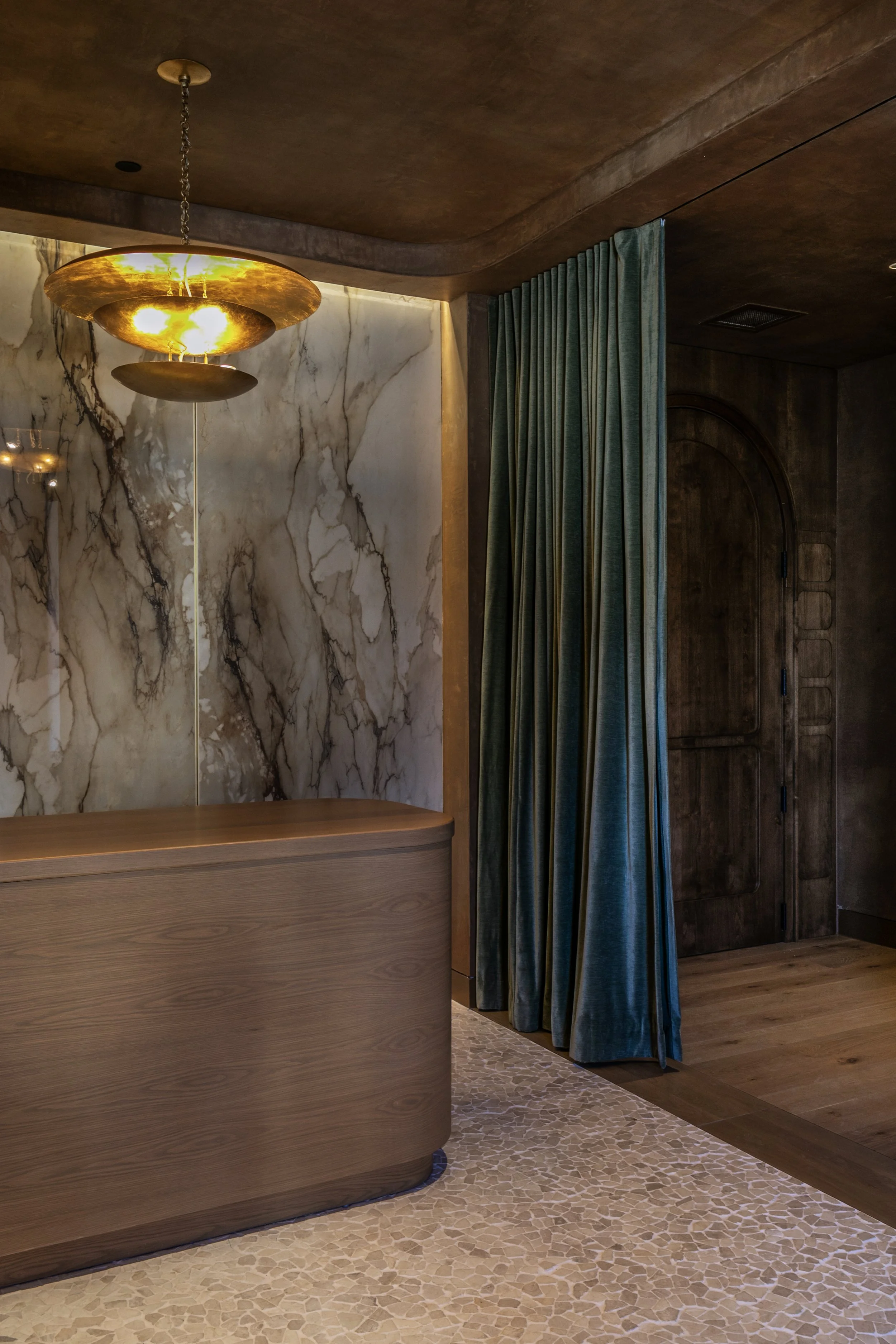 Interior of a room with a marble wall, a modern gold-colored pendant light, a wooden curved reception desk, teal curtains, a wooden door, and a pebble tile floor.