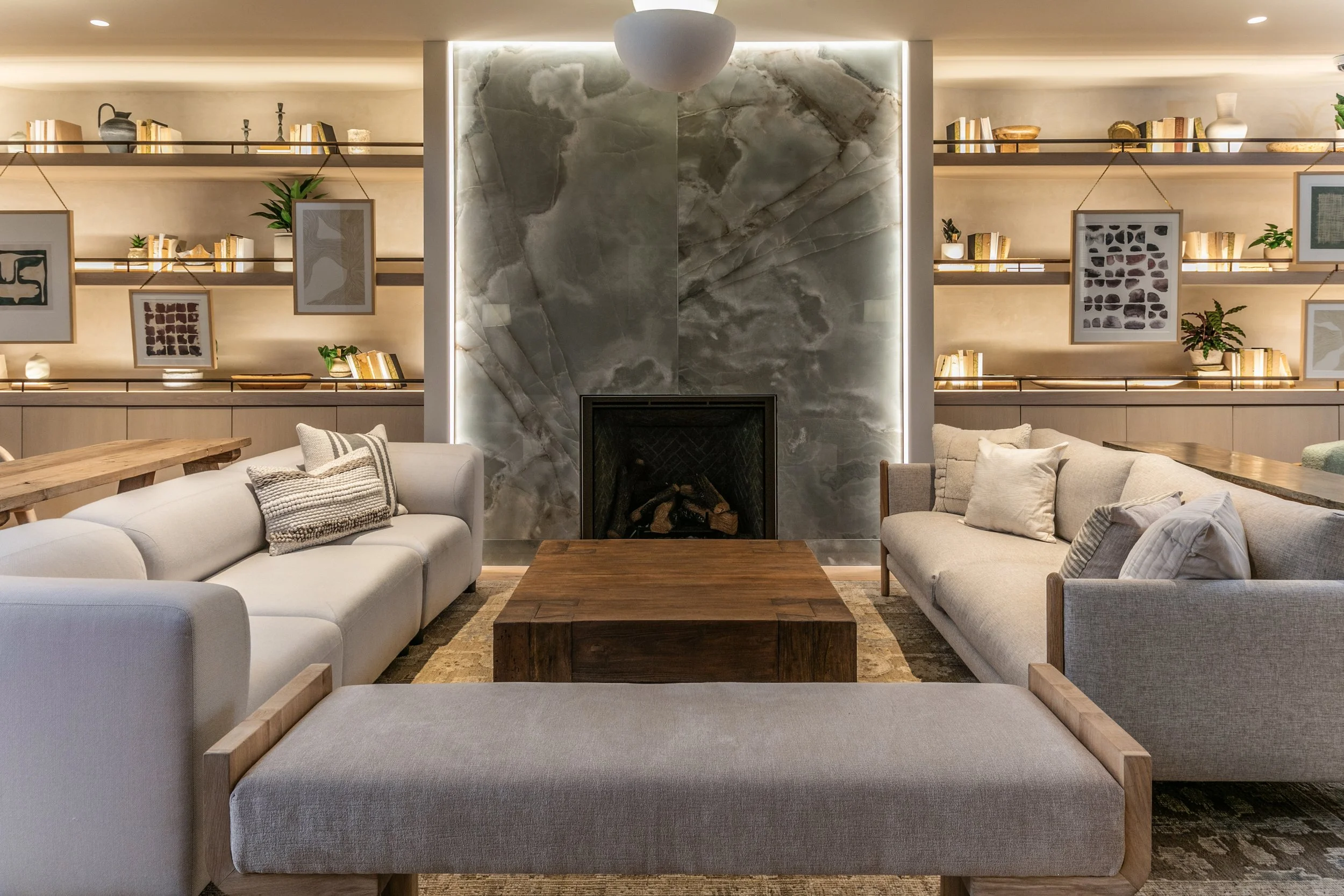 Living room with two beige sofas, a wooden coffee table, a marble fireplace, and built-in shelves with books, artwork, and decorative items.