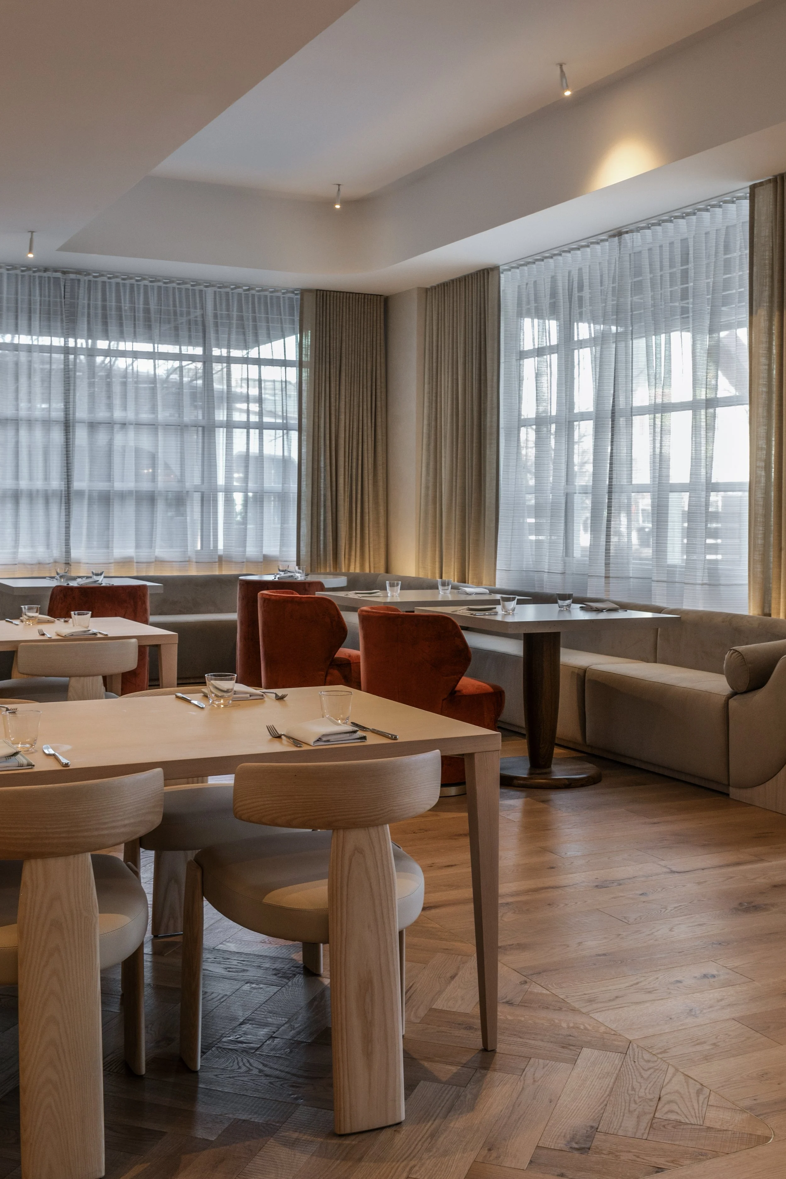 Empty restaurant dining area with wooden tables, beige and red chairs, and large windows with sheer curtains.