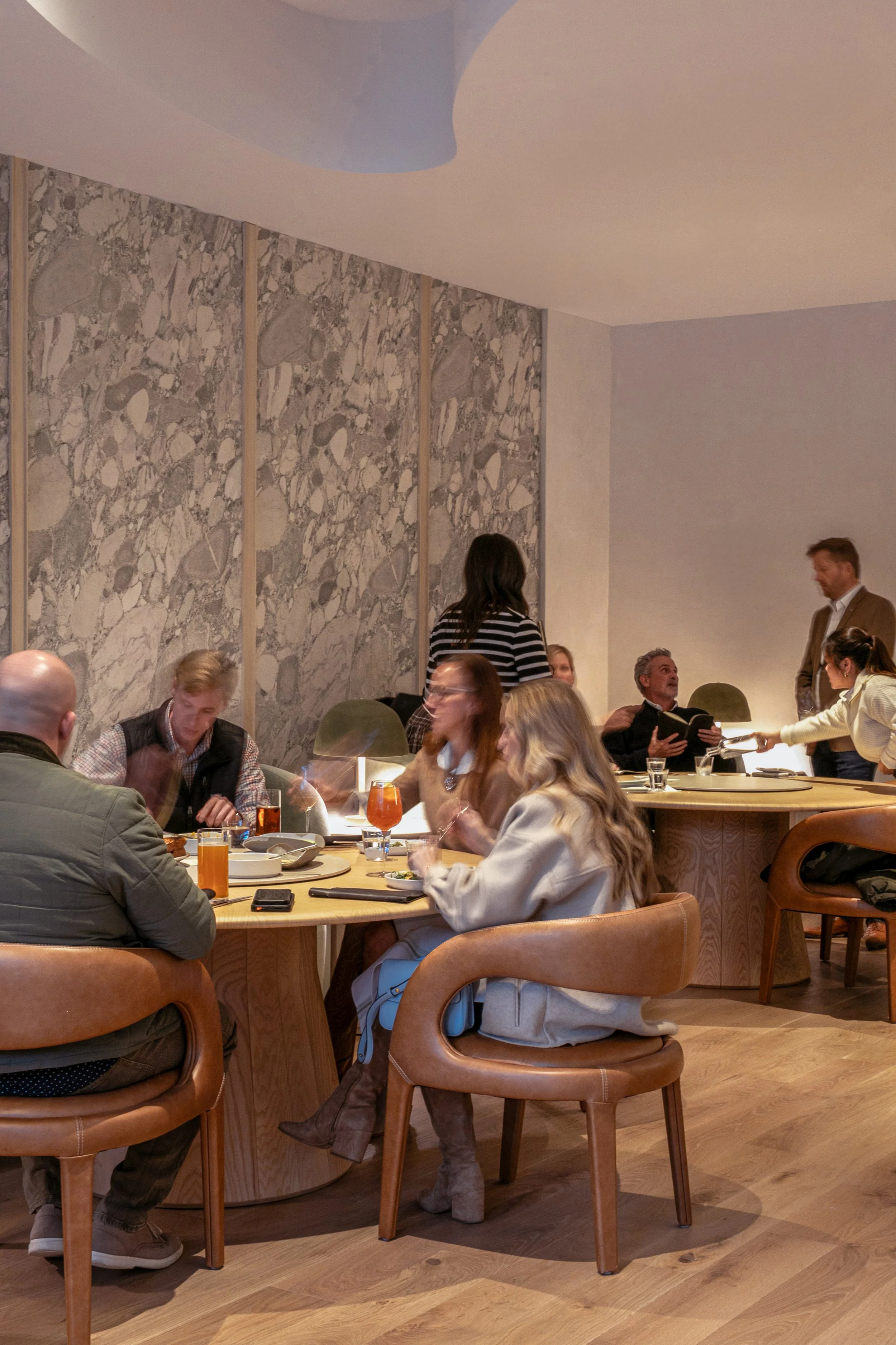 People dining at a restaurant table with drinks and food, with some interaction among the guests.