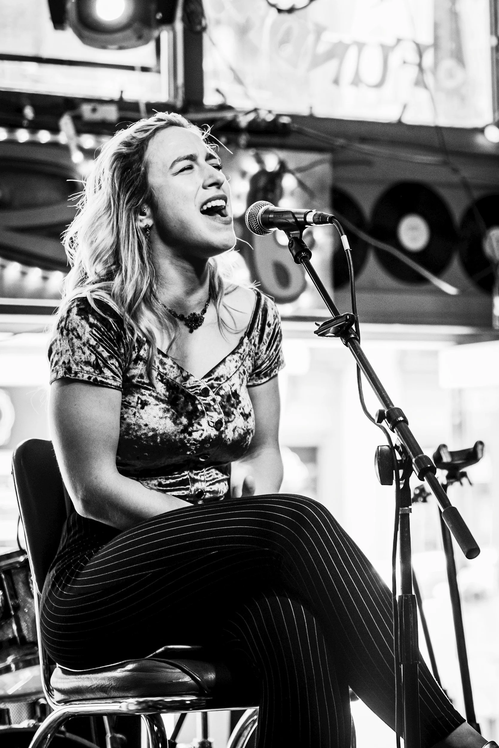 Black and white photo of a blond girl sitting on a stage singing into a microphone.
