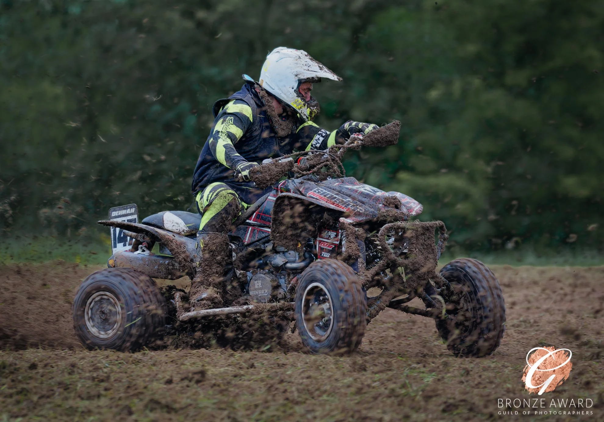 A person riding a dirt bike through a muddy off-road trail, wearing a white helmet and motocross gear.