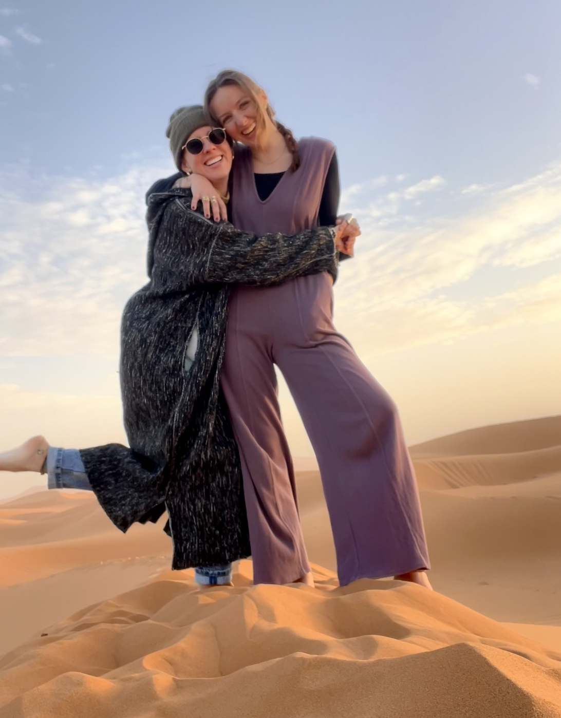 Two women are embracing and smiling in a desert landscape during sunset, with sand dunes and a partly cloudy sky in the background.