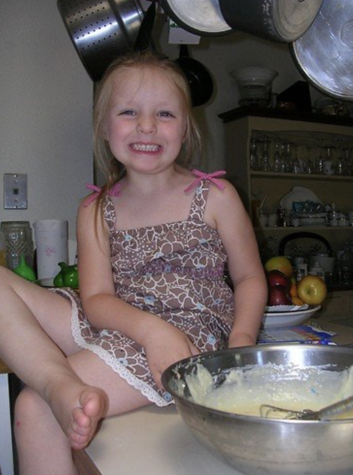 A young girl with blonde hair, wearing a brown dress with a pink ribbon on the shoulder, is sitting on a kitchen counter and smiling at the camera. In front of her is a metal mixing bowl with a spatula inside, containing a creamy pale yellow mixture. Behind her are kitchen shelves with glassware, and a bowl of apples.