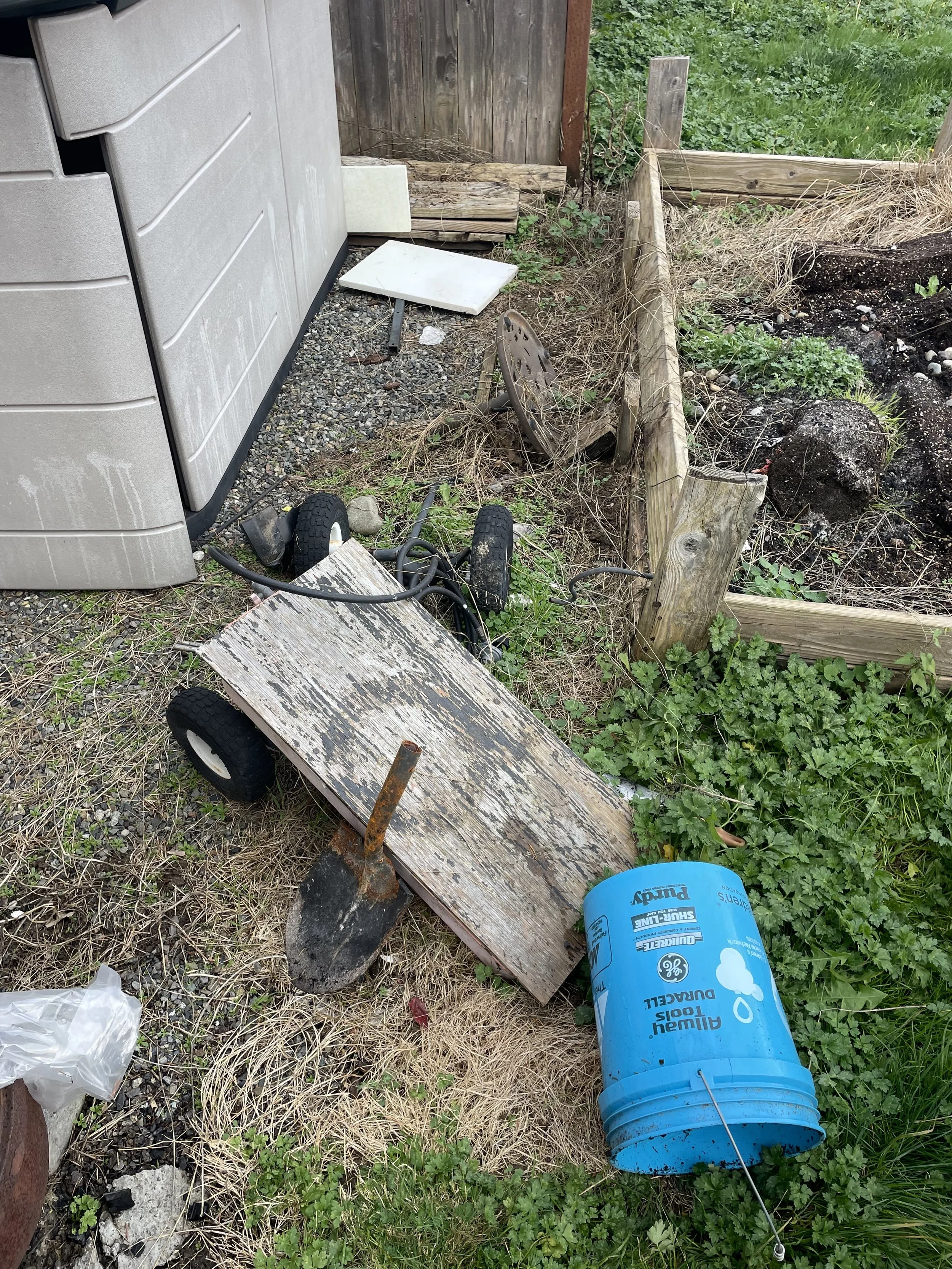 Side yard corner filled with scrap wood, tools, buckets, and overgrown weeds prior to removal.