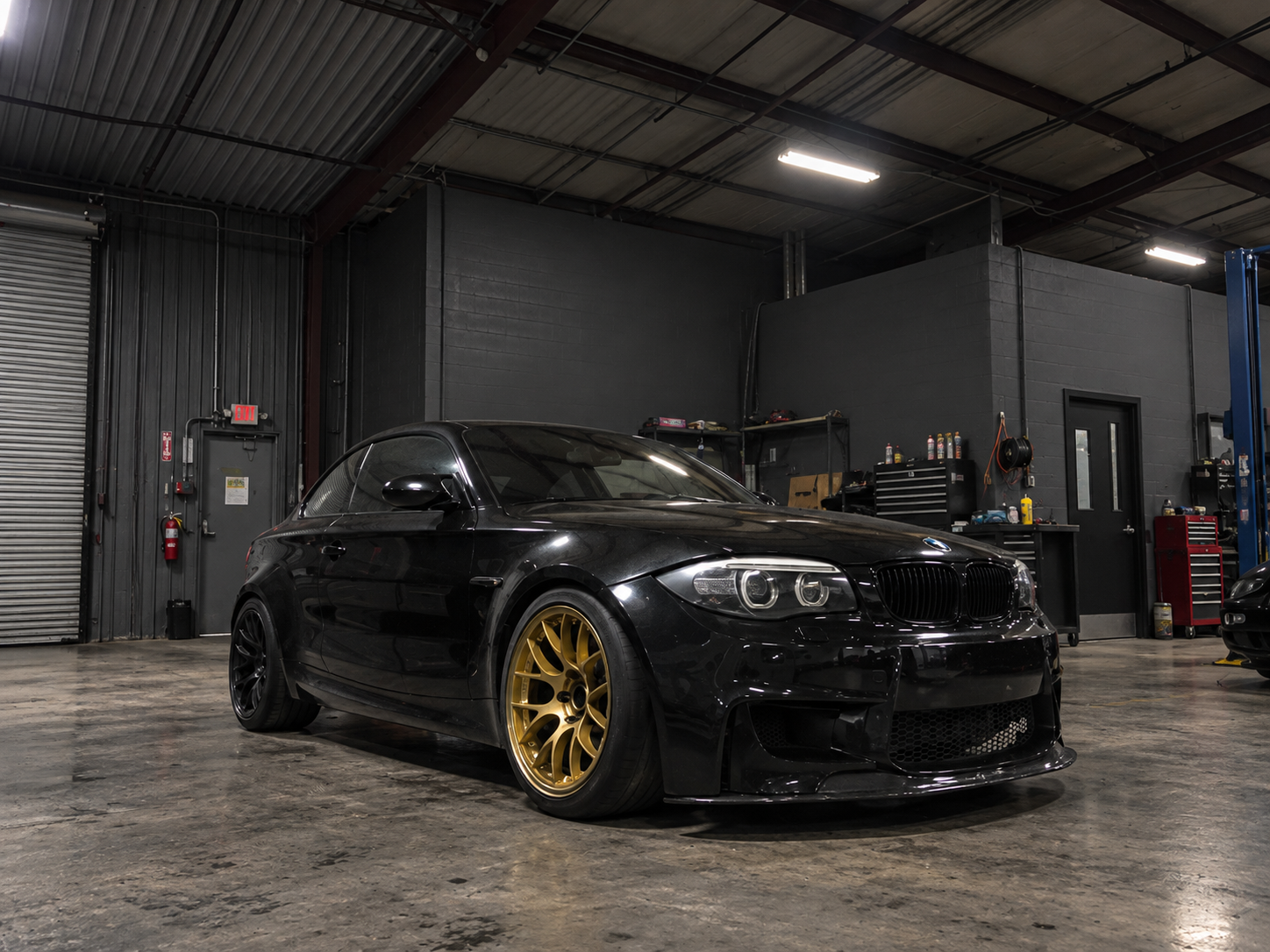 A black BMW 1M coupe with gold wheels parked inside a warehouse or garage with a concrete floor, white walls, and a beige ceiling with exposed beams.