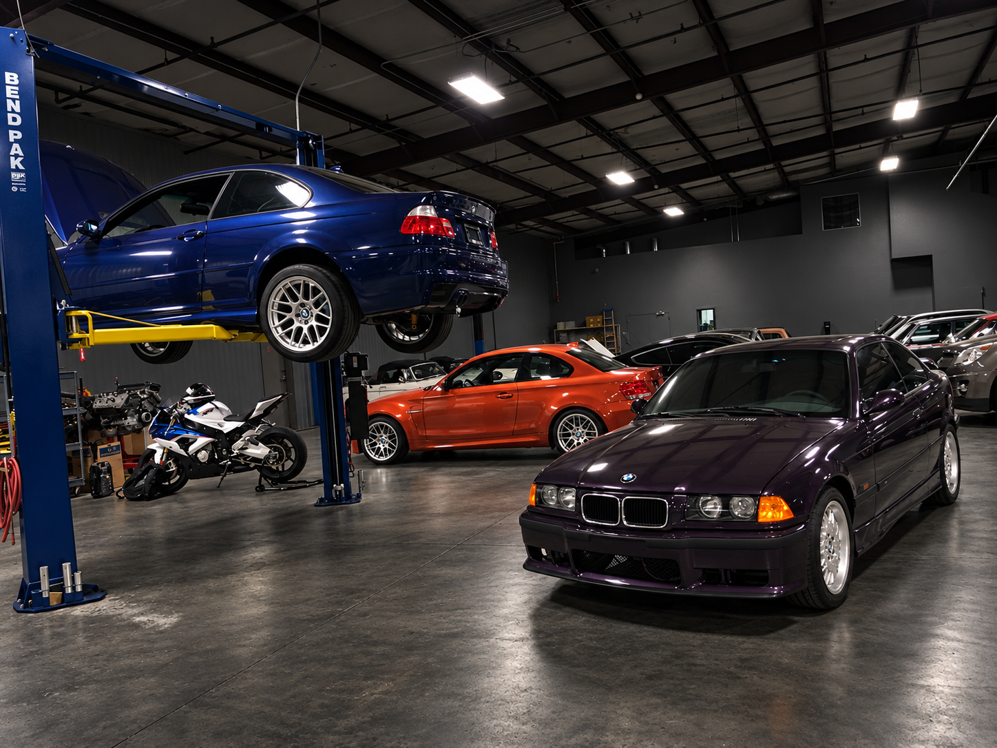 Four BMW M3 coupes lined up outside a modern charcoal garage at dusk, led by a black E46 M3 with number 13, dark wheels, and a red tow hook. Warm architectural lighting reflects across the pavement beneath a dramatic evening sky.