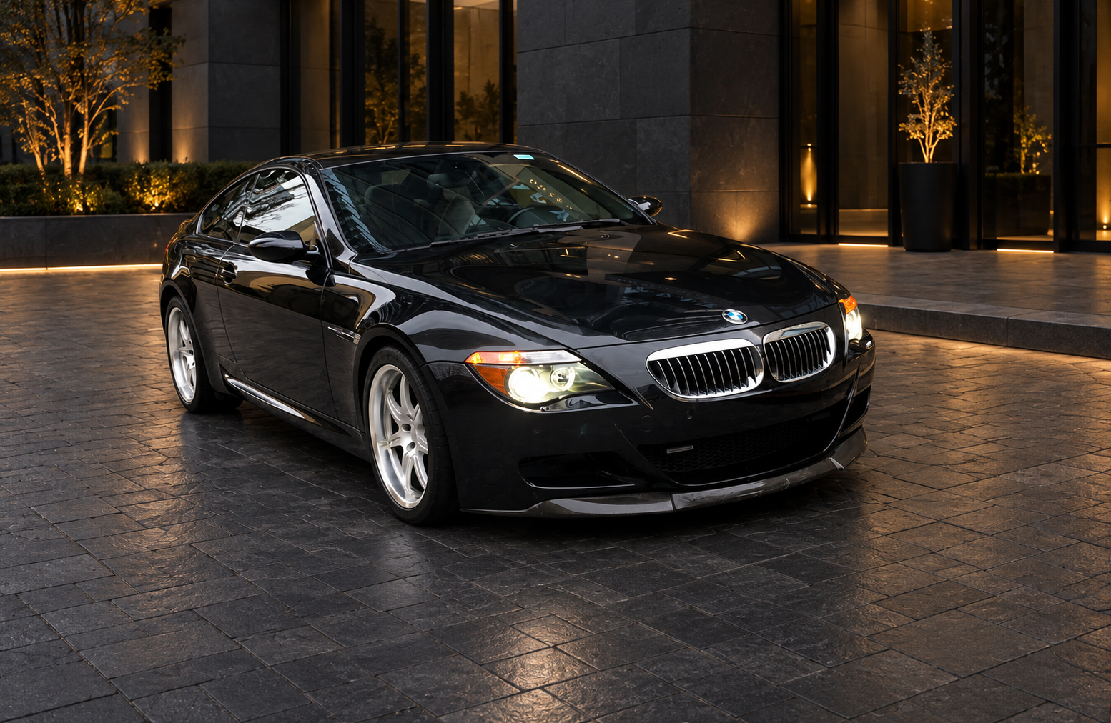 Black BMW M6 with a Dinan 5.7L stroker V10, silver DINAN wheels, and illuminated headlights, parked outside a sleek modern building with charcoal stone, tinted glass, and warm architectural lighting.