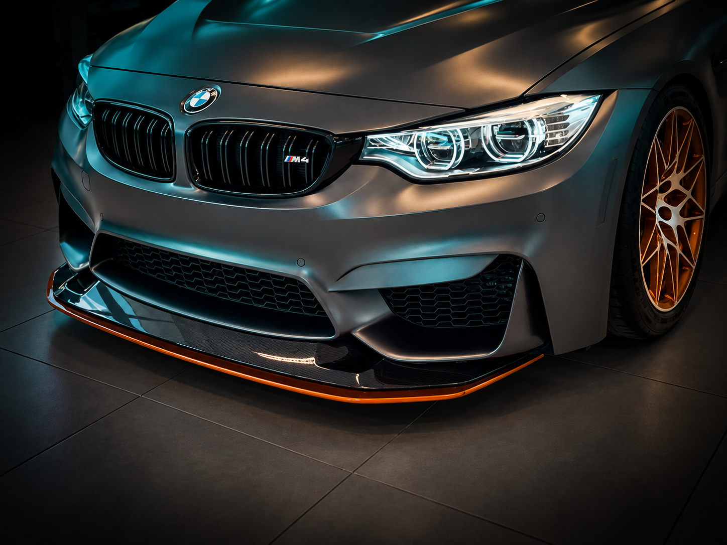 Close-up of a high-performance gray BMW M4 sports car with a black and blue kidney grille, LED headlights, orange accents on the front splitter, and distinctive gold alloy wheels, parked indoors with reflections on its surface.
