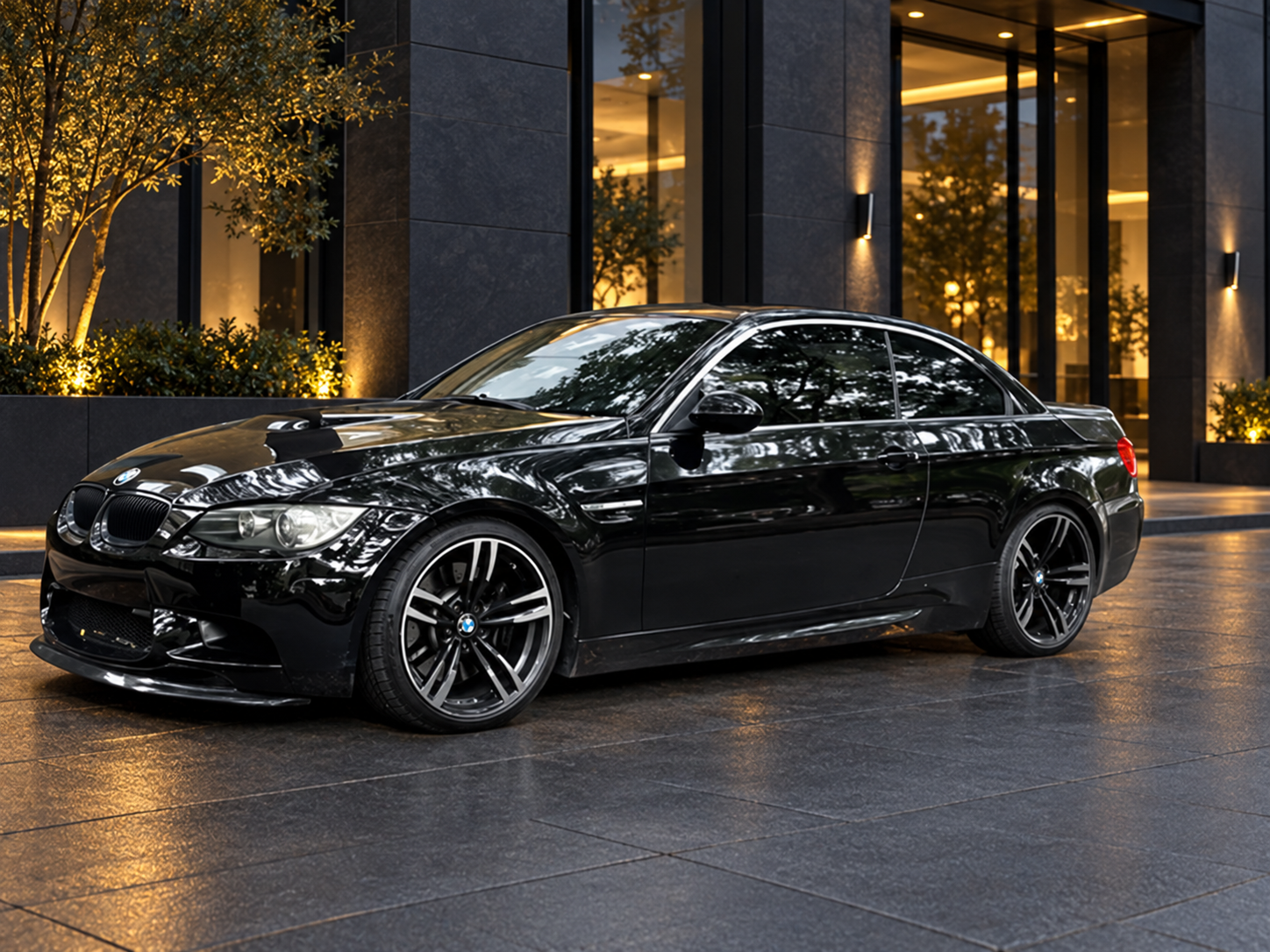 A black BMW E93 M3 convertible car parked on the street in front of a brick building with windows, surrounded by trees and fallen autumn leaves.