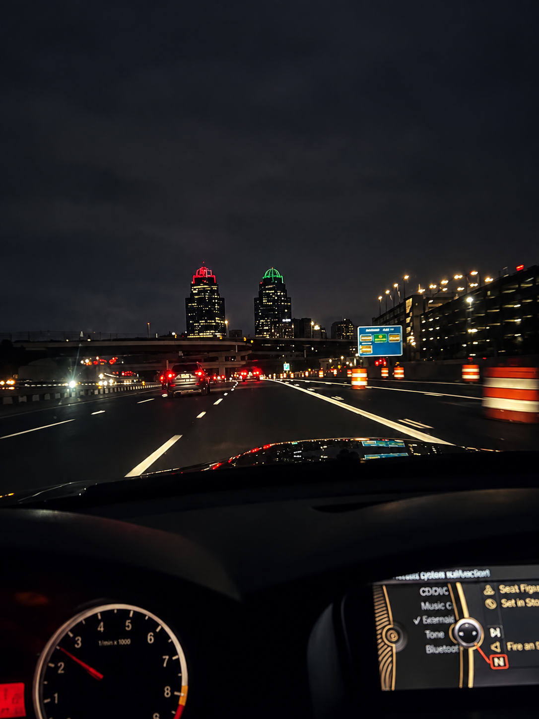 Nighttime view from inside a car on a highway with two tall buildings in the distance, one with red lights at the top and the other with green lights, and traffic cones along the side of the road. A GPS navigation screen is partially visible on the d