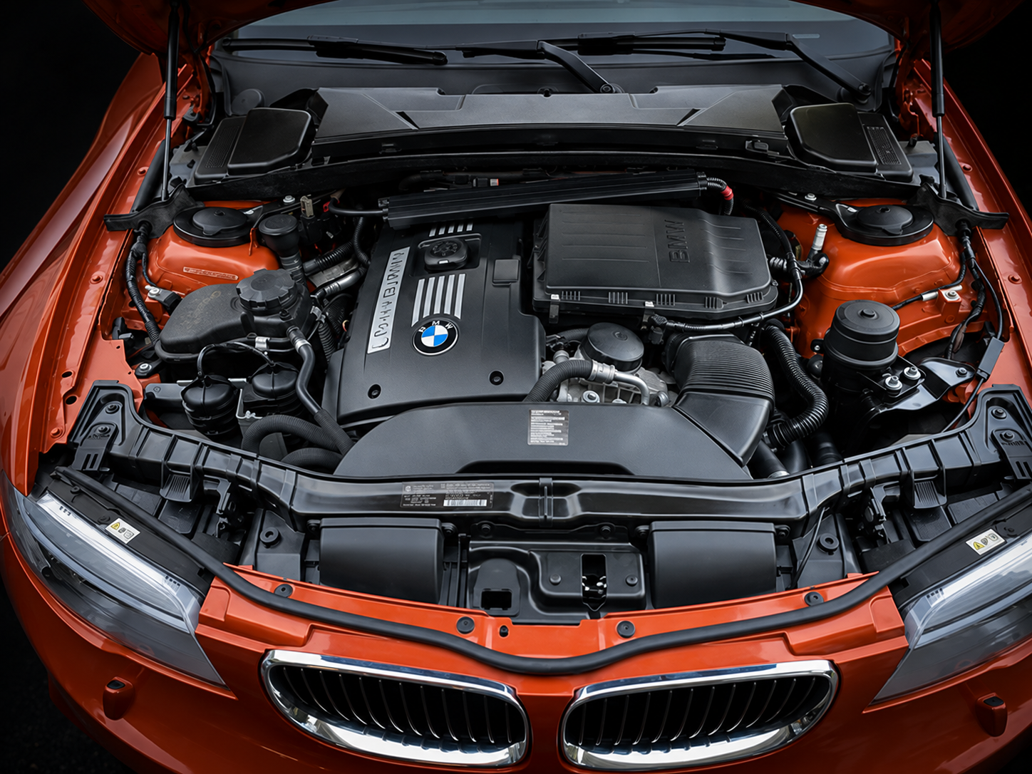 Front view of a red BMW car with its hood open, revealing the engine bay with a black engine cover, various hoses, and components.