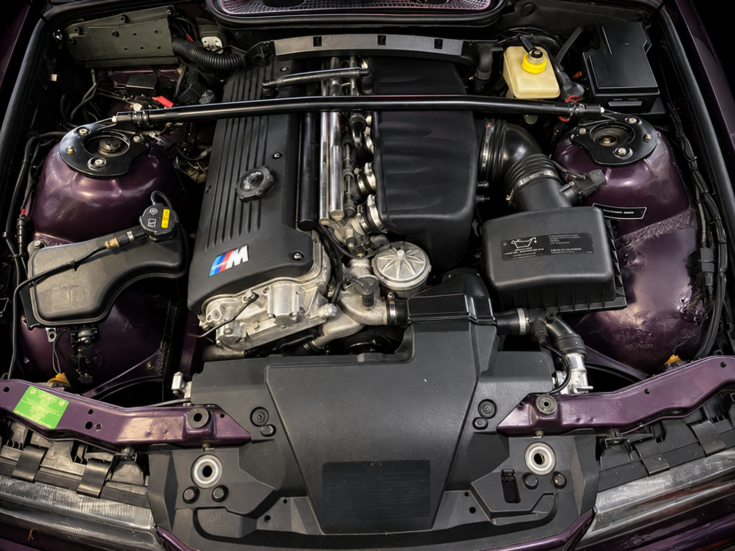The image shows the engine bay of an S54 swapped E36 M3, featuring the S54 engine various mechanical components, hoses, and parts of the engine.