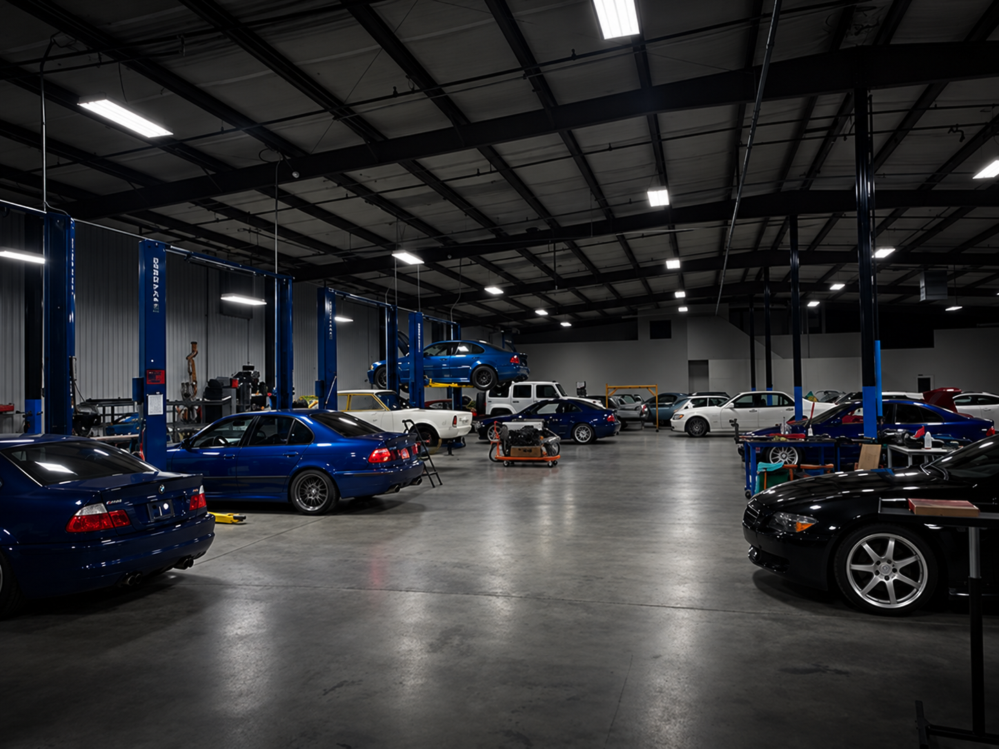 Inside a spacious automotive repair shop with a high ceiling, numerous cars are parked, including a blue sports car elevated on a lift, various tools and equipment are visible, and overhead lighting illuminates the area.