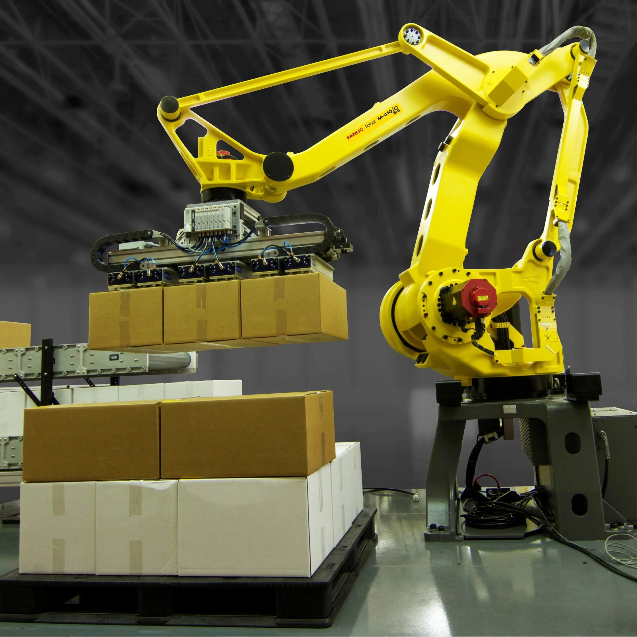 Yellow robotic arm with the label 'FANUC Robot M-410iC' assembling cardboard boxes in an industrial setting.