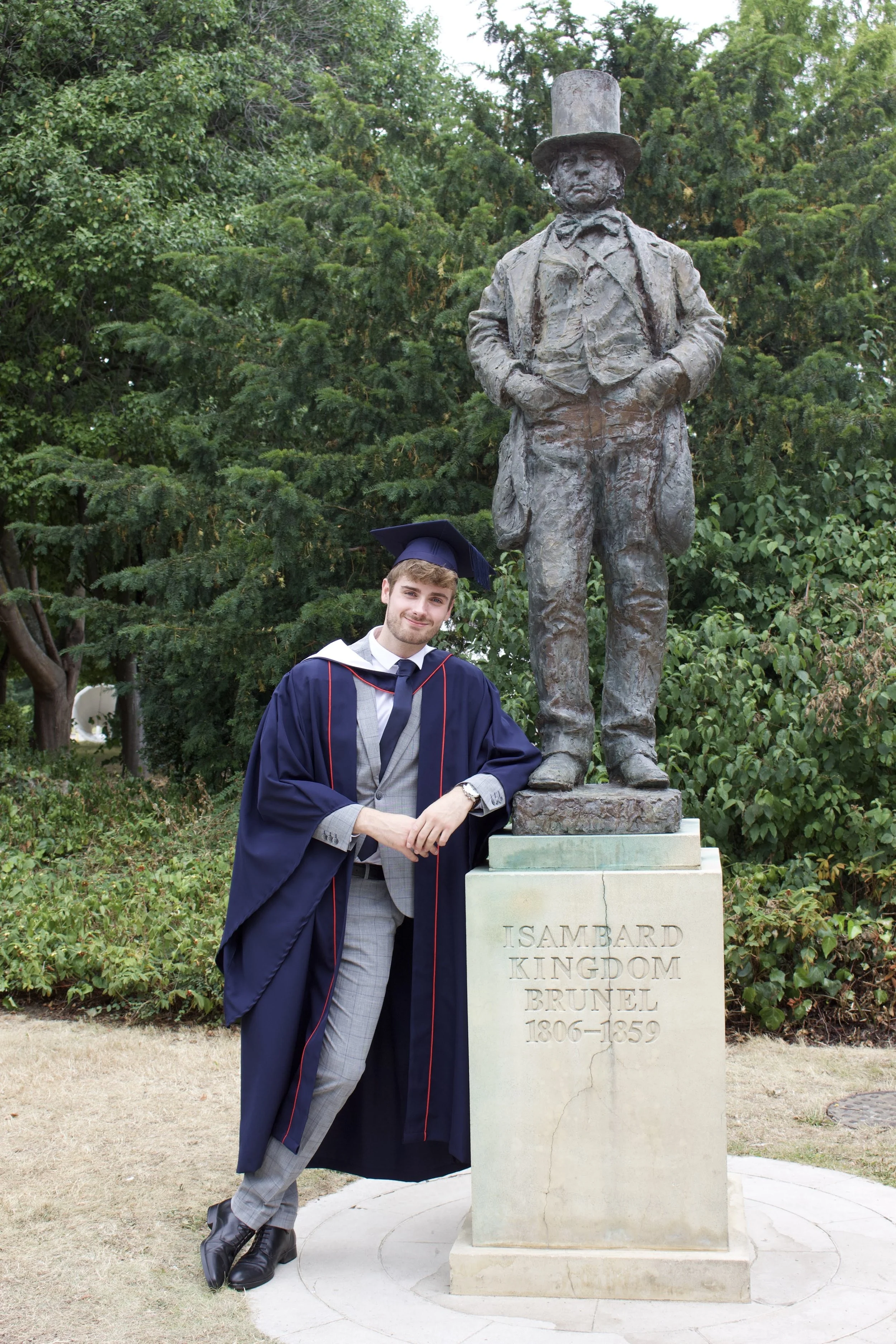 Man in a red cycling jersey and helmet standing in front of a scenic coastal background with a clear blue sky, turquoise sea, and rocky cliffs.