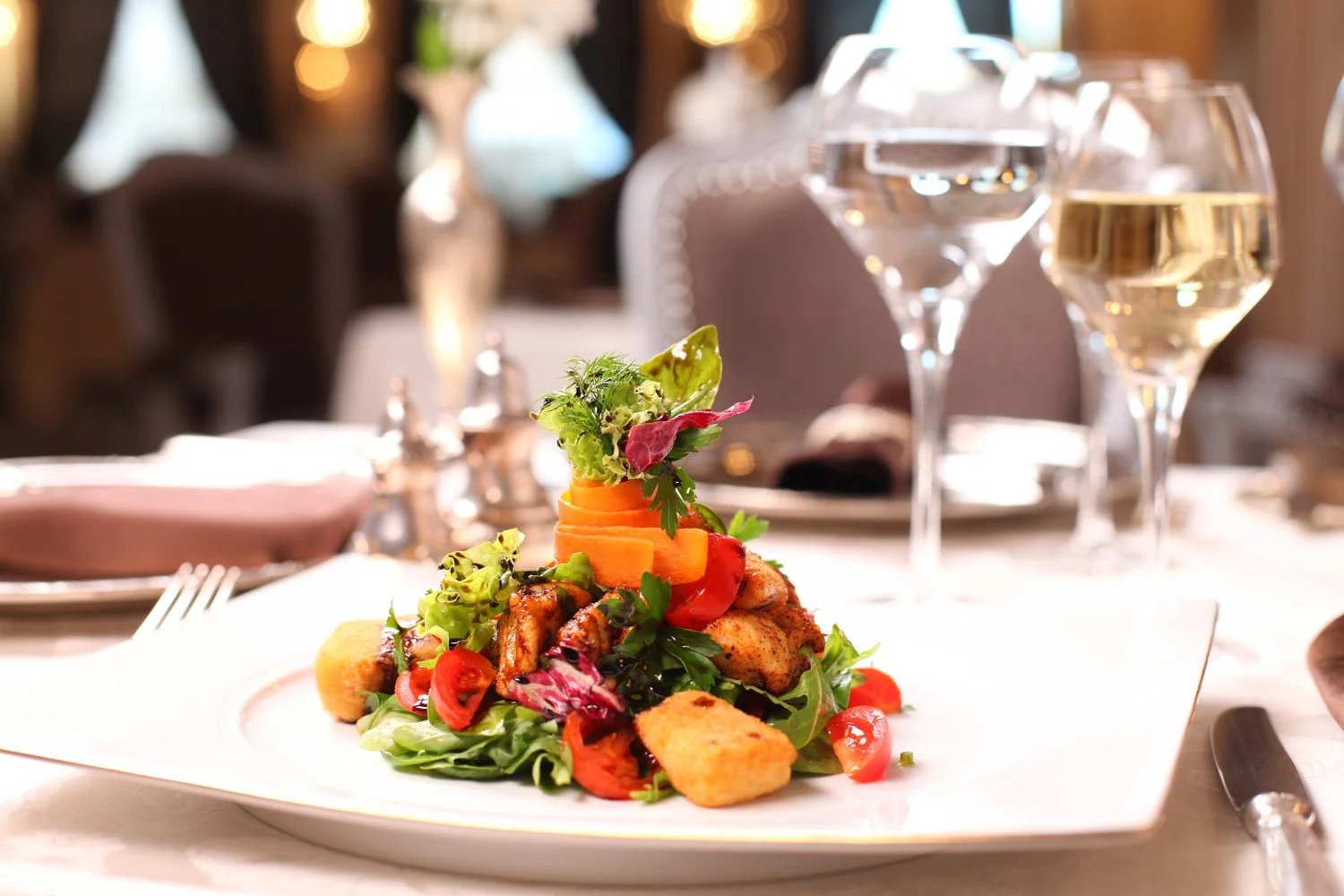 Ein Teller mit Salat, gegrilltem Hühnchen, Tomaten, Karotten, und Croûtons auf einem festlich gedeckten Tisch mit Weingläsern und Besteck in einem Restaurant.