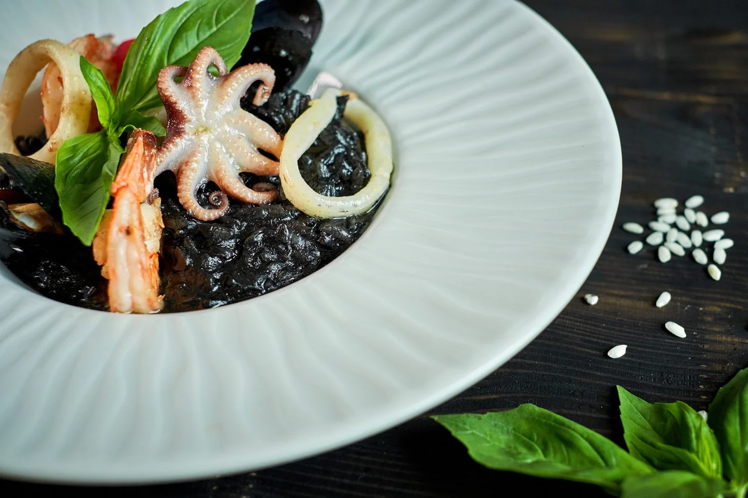 Schwarz gefärbter Tintenfisch in einer weißen Schale mit Muscheln, Zwiebelringen und Basilikum, auf einem dunklen Holztisch mit weißen Sesamsamen und Basilikumblättern