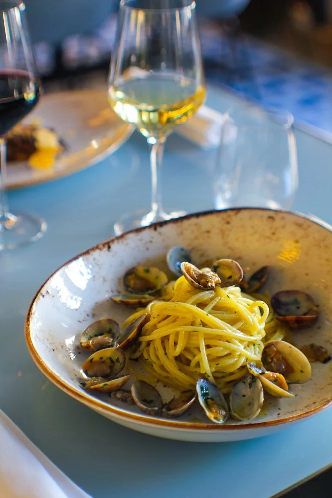 Eine Schüssel mit Spaghetti mit Muscheln an einem Esstisch, im Hintergrund Wein und weitere Speisen.