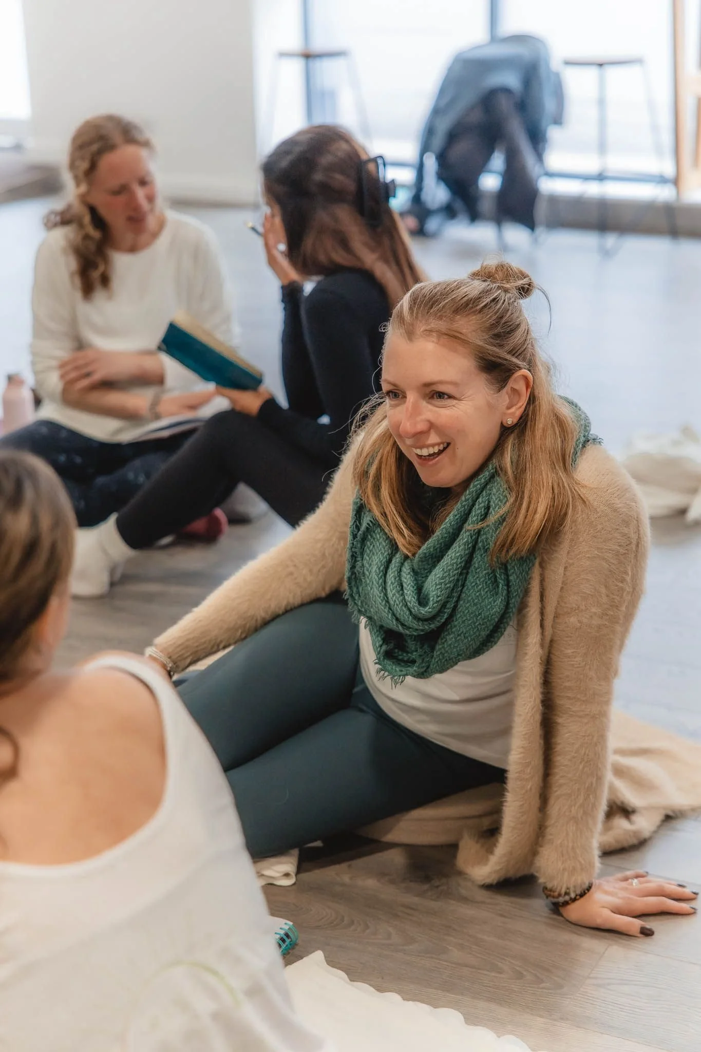Women sitting on the floor in a circle during a discussion or class, with one woman smiling and others engaged, in a bright room with large windows.