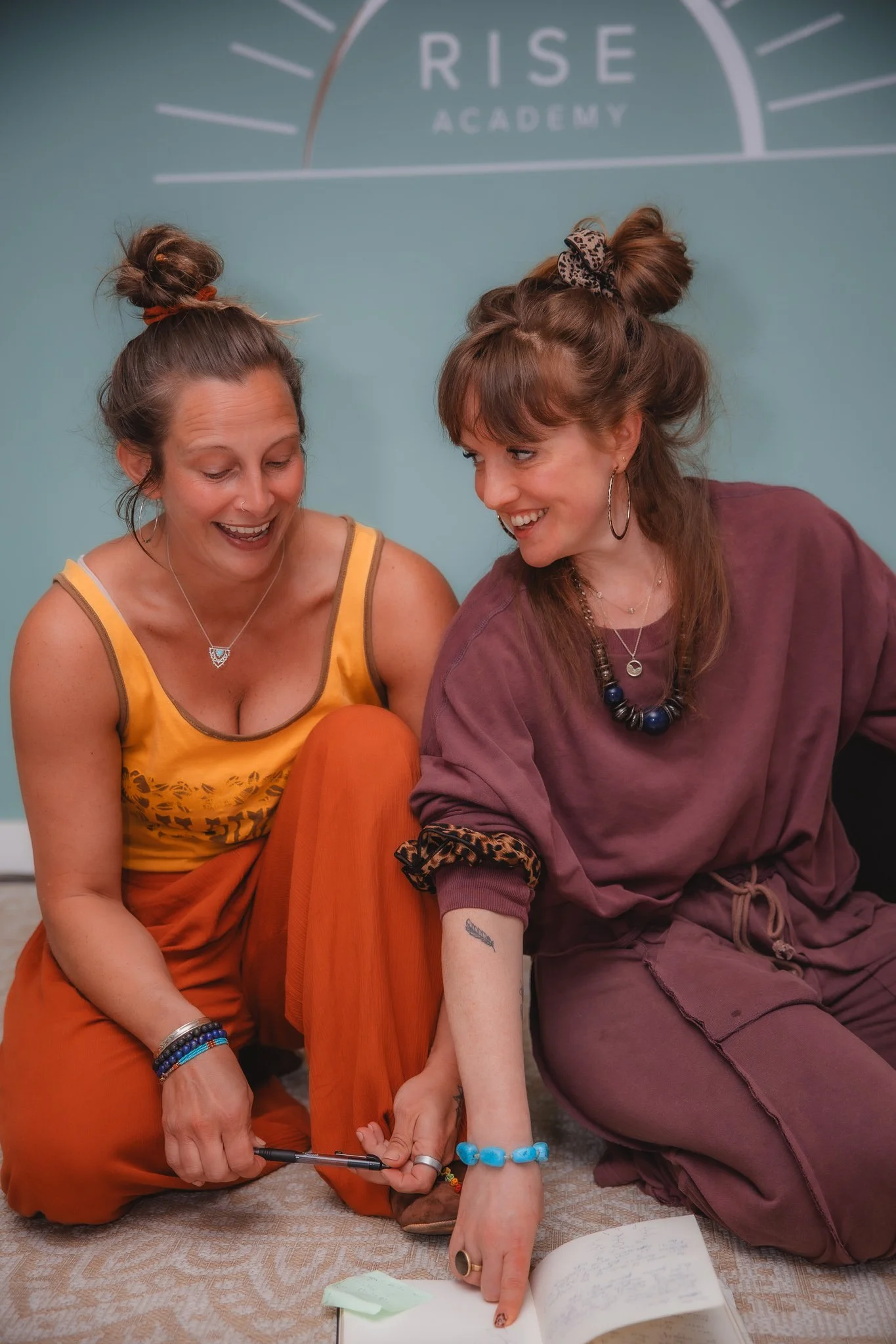 Two women smiling happily, standing close with one arm around each other, wearing matching dark green hoodies with 'RISE' logo, against a light green wall with white sunburst design.