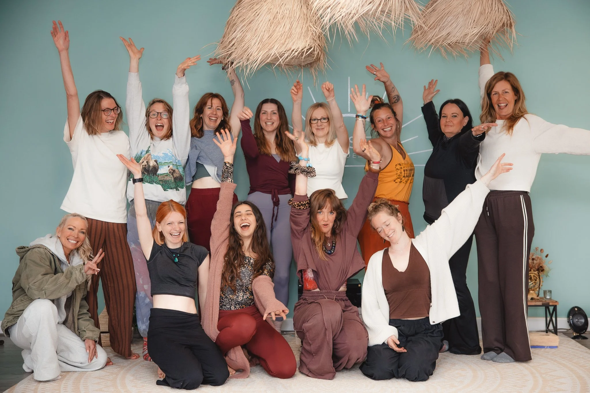 Group of women of diverse ages and ethnicities smiling and raising their arms indoors in front of a teal wall with a thatched roof decoration.