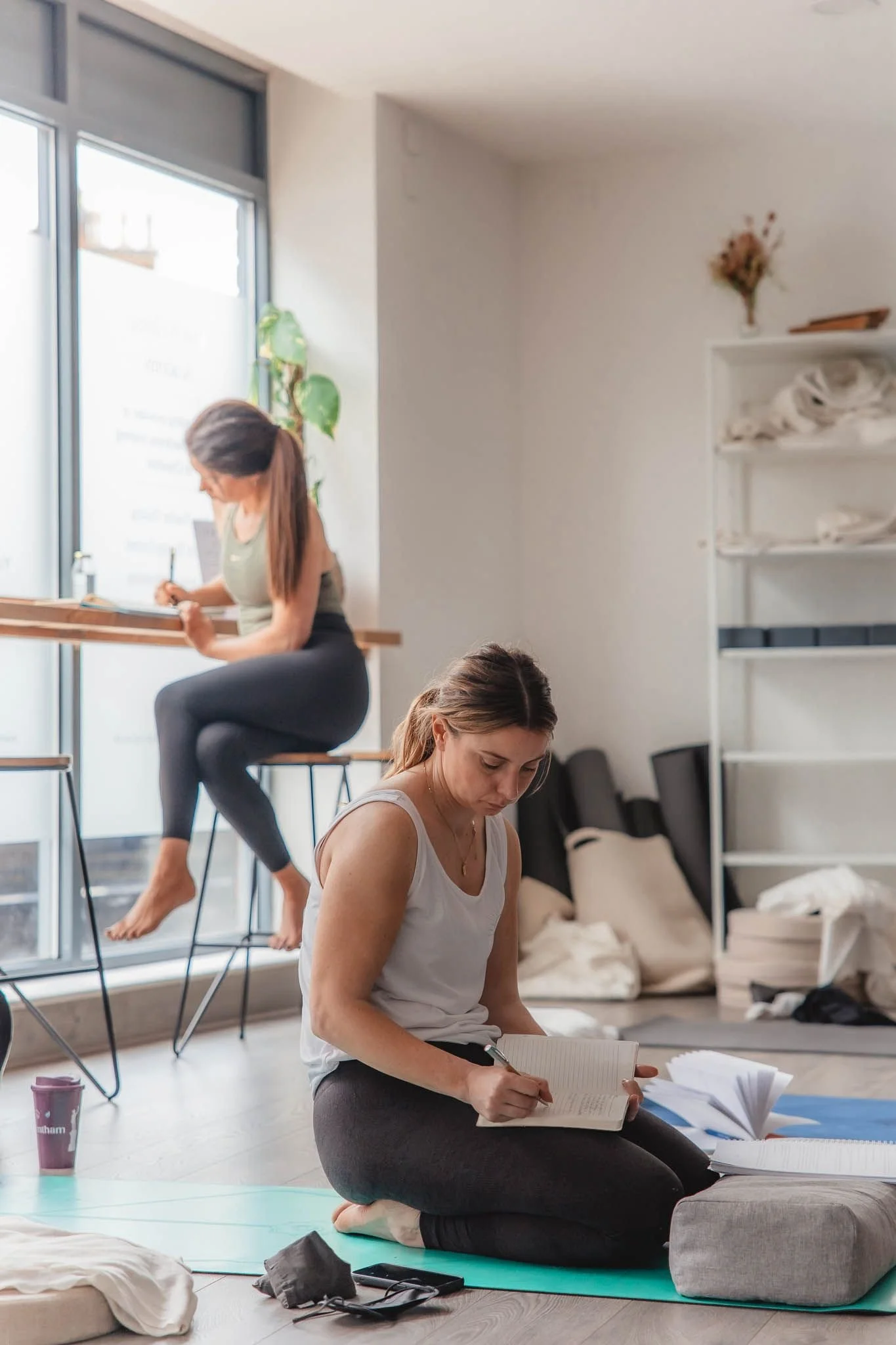 Two women practicing yoga and meditation in a bright room with large windows, books, and supplies around.