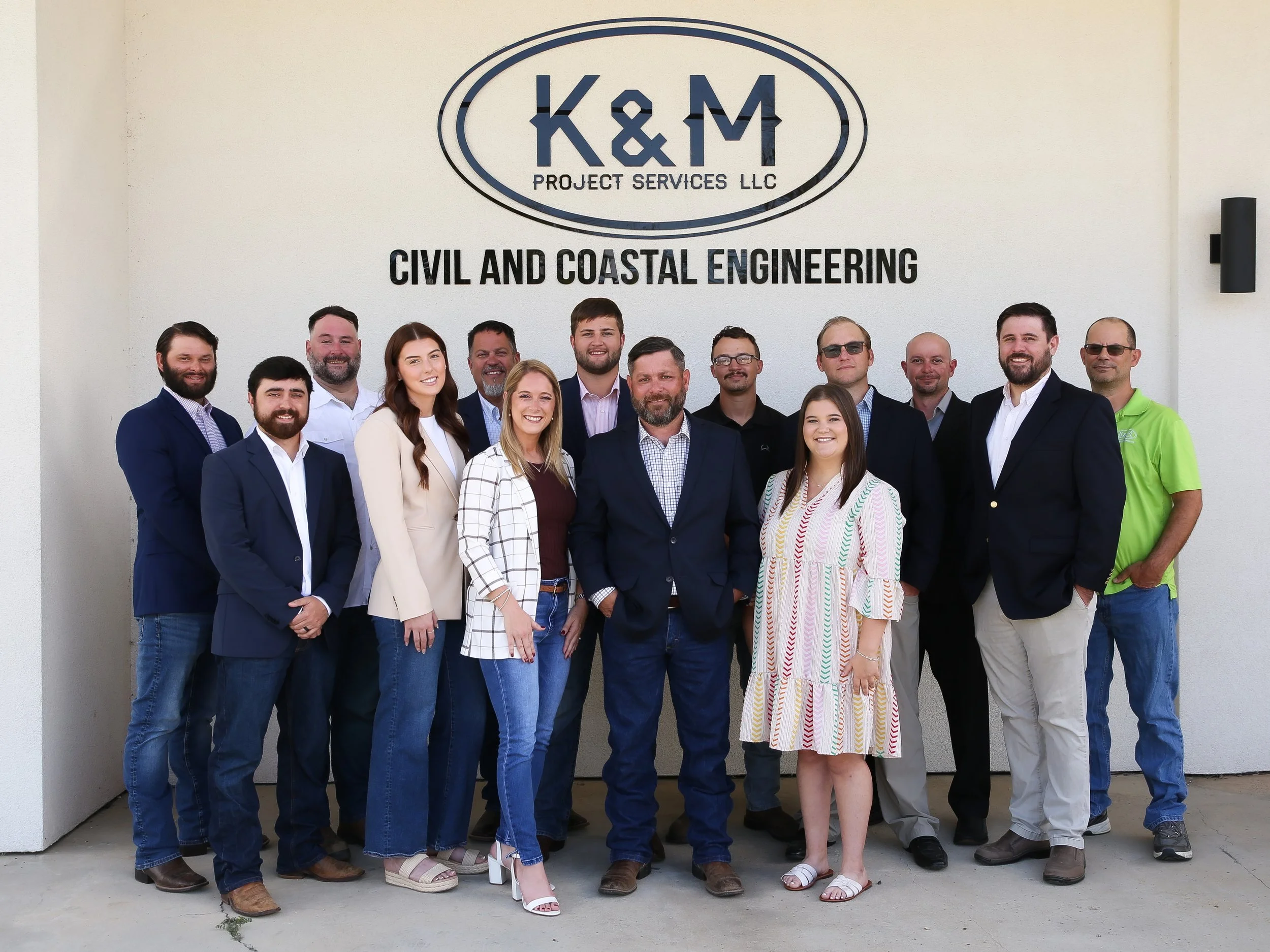 A group of people standing in front of a wall with the logo and name of K&M Project Services LLC, specializing in civil and coastal engineering.