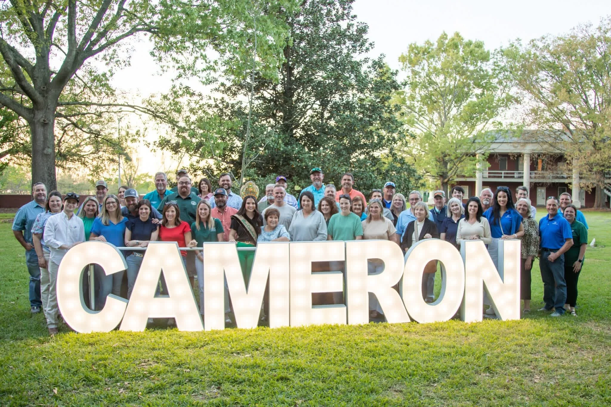 A large group of people standing outdoors on a grassy area, posing for a photo behind large illuminated letters spelling 'CAVEXPORT'. There are trees and a building in the background, and the photo is taken during daylight.