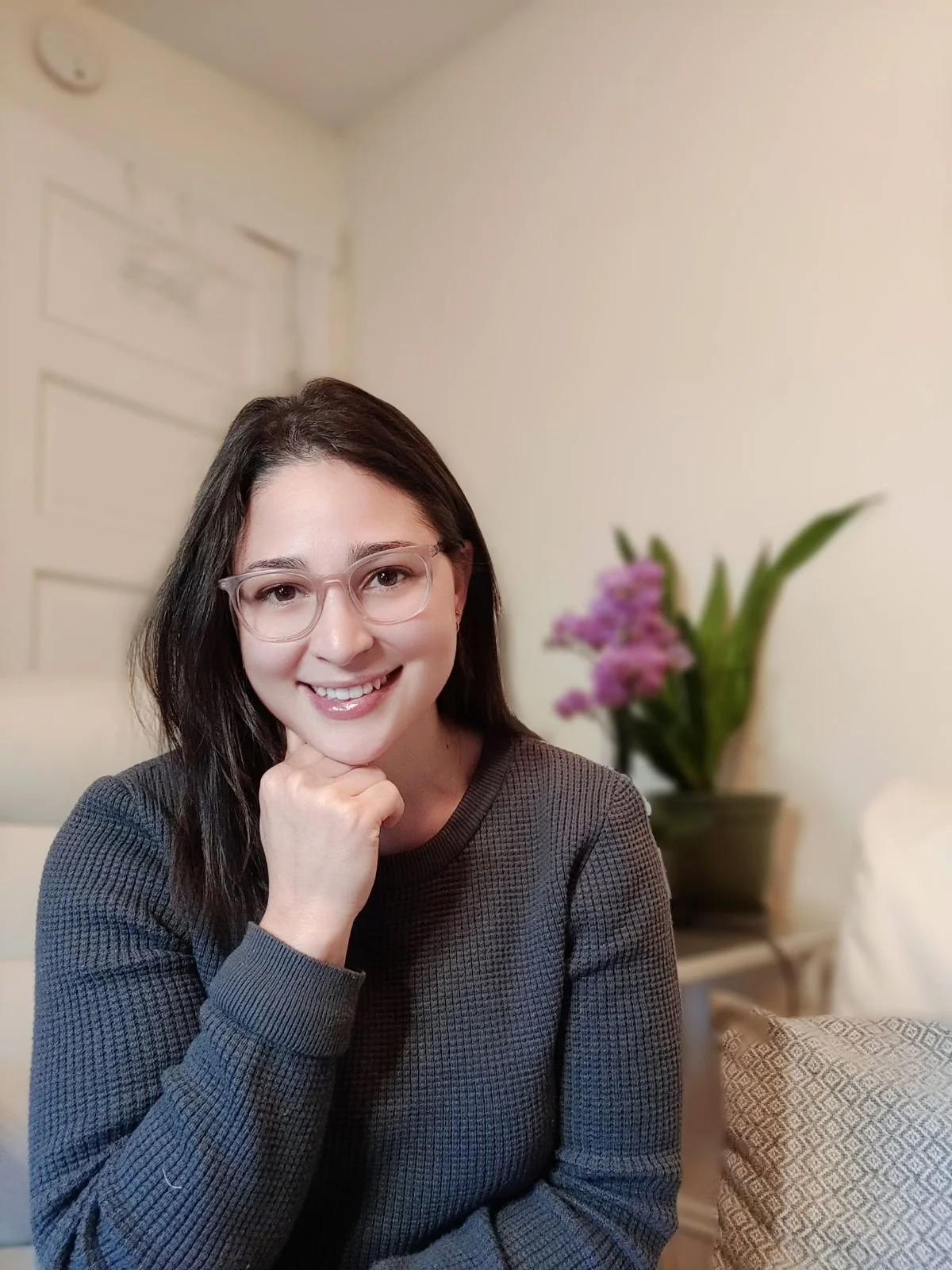 Person wearing glasses and a blue sweater, smiling indoors with green plant and framed picture in background.