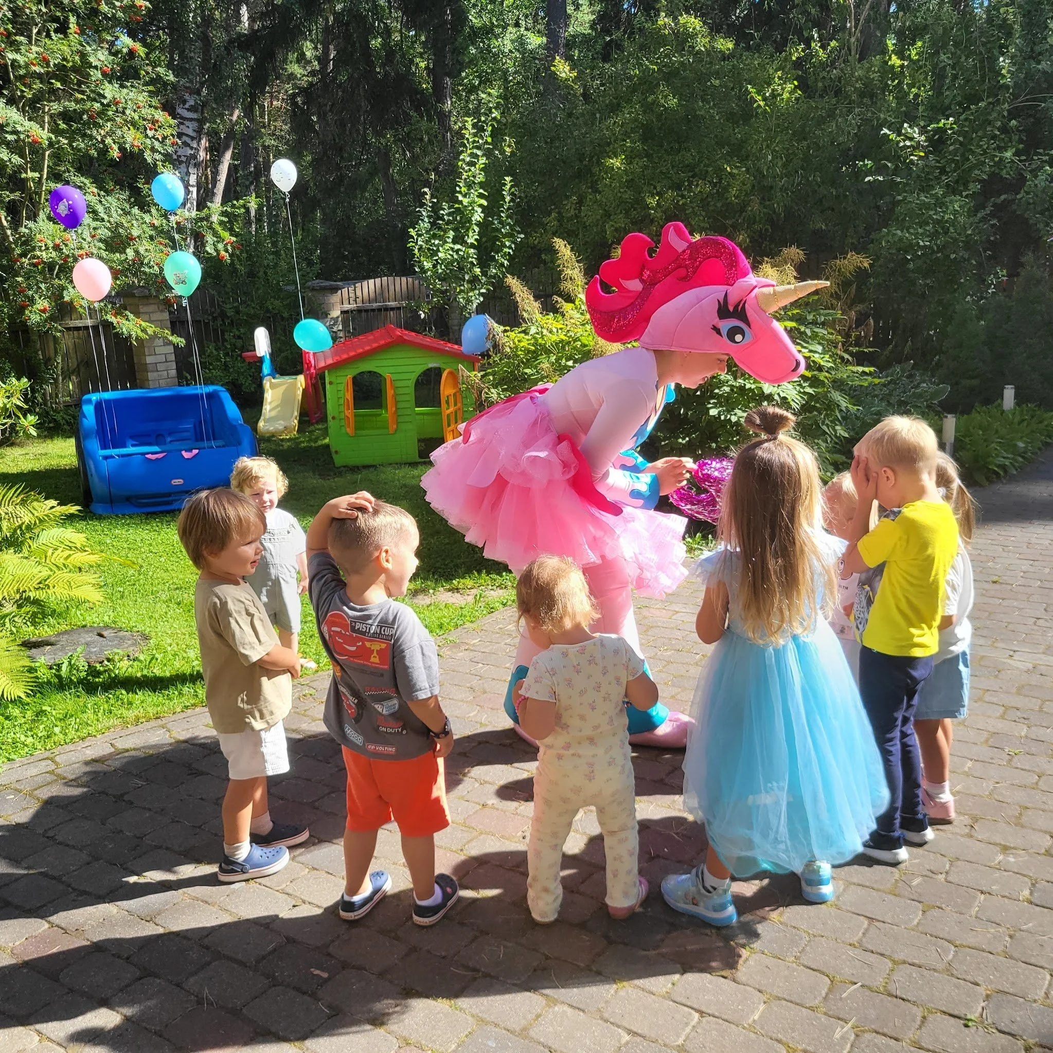A children's birthday party outdoors with a person dressed in a pink unicorn costume entertaining a group of young kids. The scene features a green backyard with a playhouse, balloons, and sunny weather.