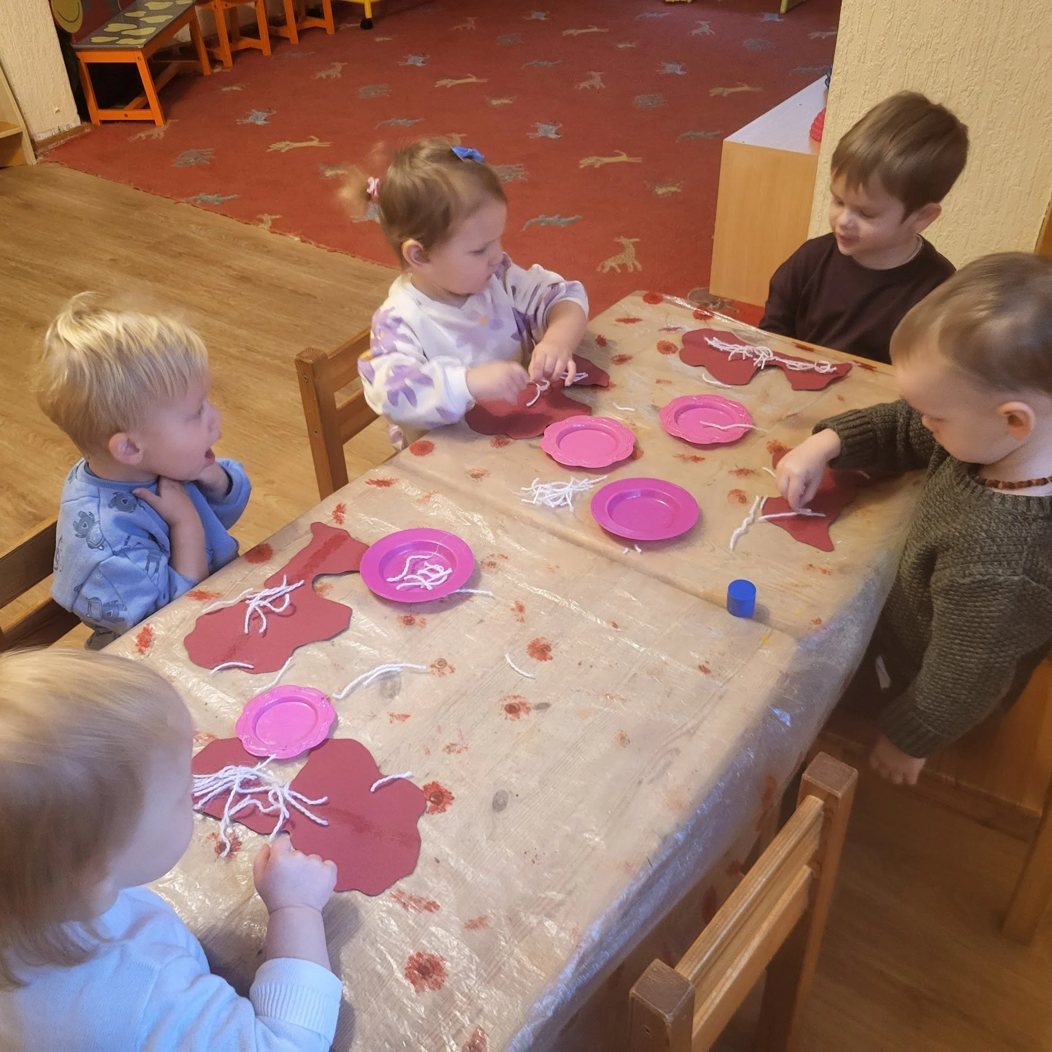 Five young children sitting at a table making crafts with red paper cutouts, white yarn, and pink paper plates.