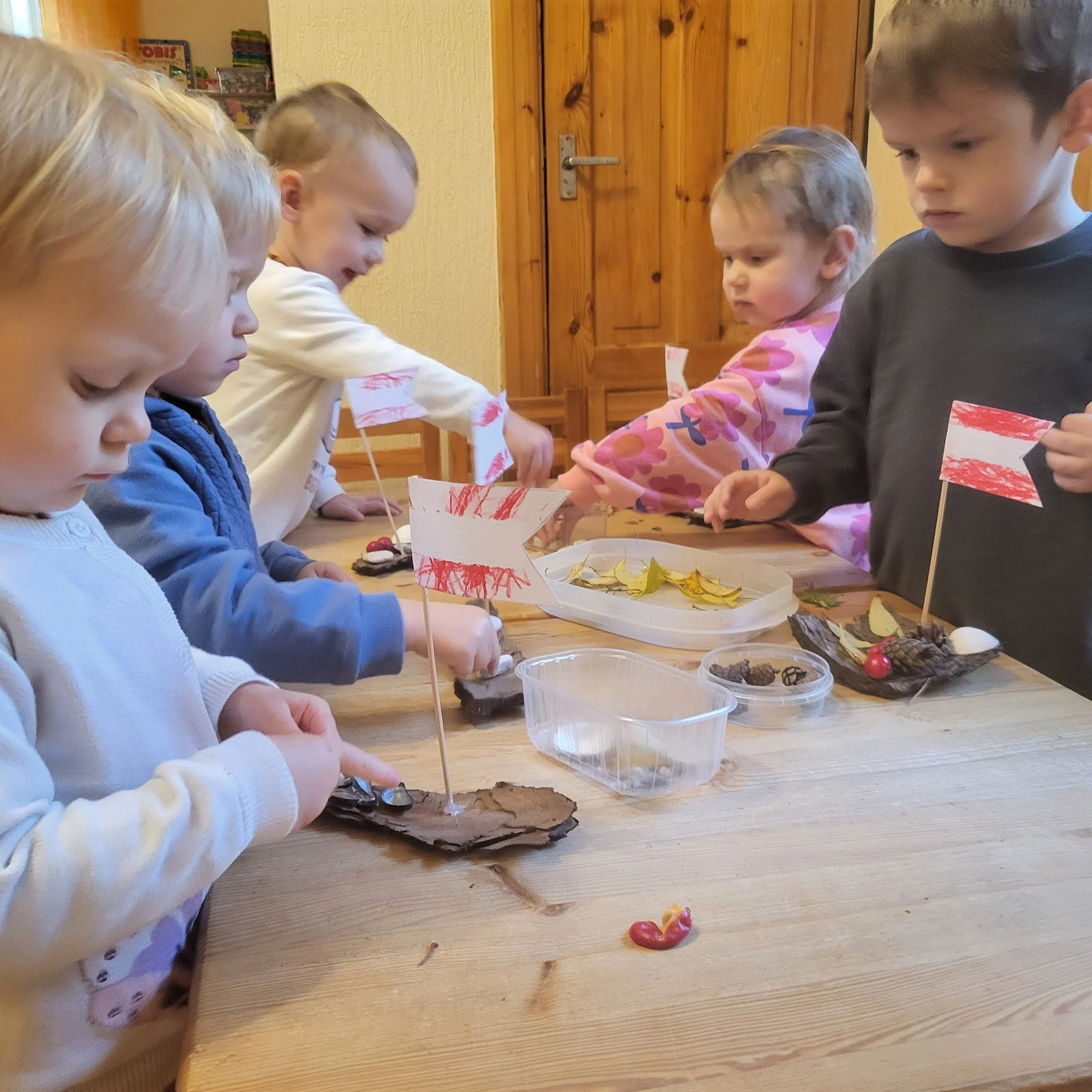 Six young children standing around a wooden table, engaged in a craft project, with small paper flags on sticks, leaves, berries, and other natural items on the table.